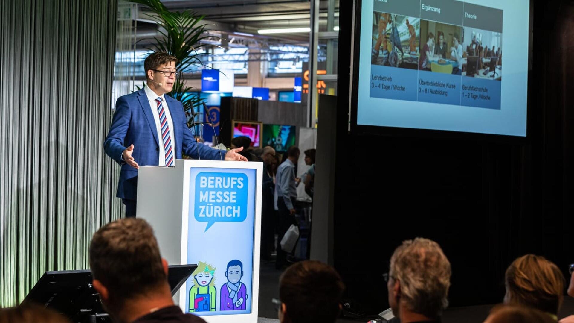 Ein Mann in einem blauen Anzug spricht an einem Podium mit der Aufschrift Berufsmesse Zürich vor einem Publikum. Auf einem großen Bildschirm wird eine Präsentation mit Bildern und deutschen Texten zum Thema Bildung oder Ausbildung gezeigt.