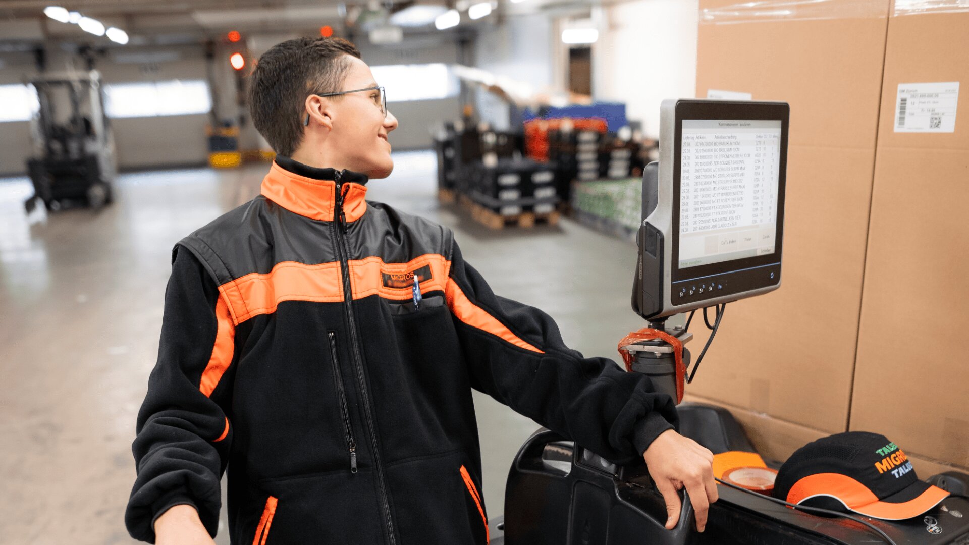 Eine Person in einer schwarz-orangenen Jacke bedient in einem Lagerhaus eine Maschine mit einem Bildschirm, auf dem Daten angezeigt werden, und blickt zur Seite, während sie neben gestapelten Kisten steht.