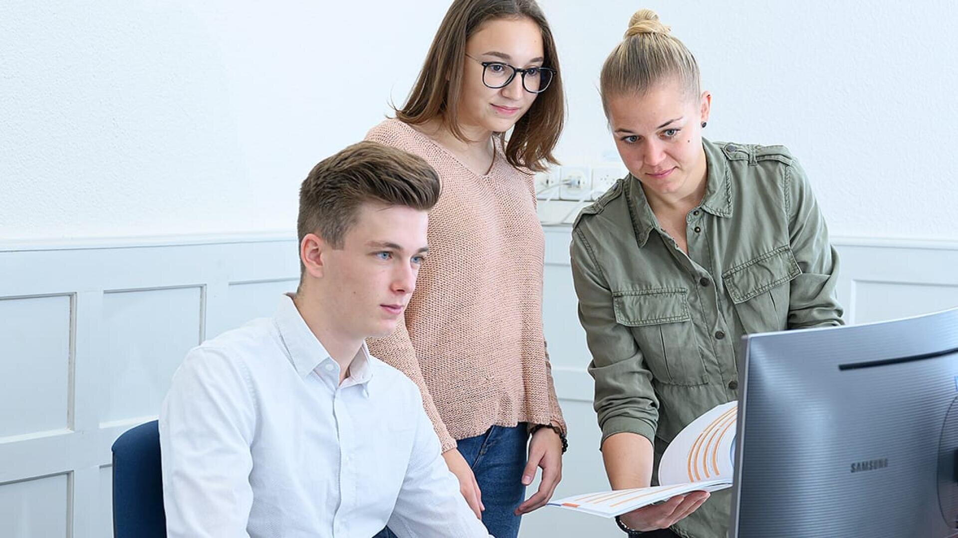 Drei junge Erwachsene arbeiten in einem Büro zusammen. Eine Person sitzt an einem Computer, während die beiden anderen daneben stehen, auf den Bildschirm schauen und gemeinsam Dokumente durchsehen.