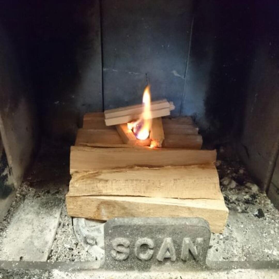 Ein kleines Feuer brennt zwischen gestapelten Holzscheiten in einem Metallkamin. Das Wort SCAN ist auf einer Metallplatte an der Vorderseite des Kamins eingeprägt.
