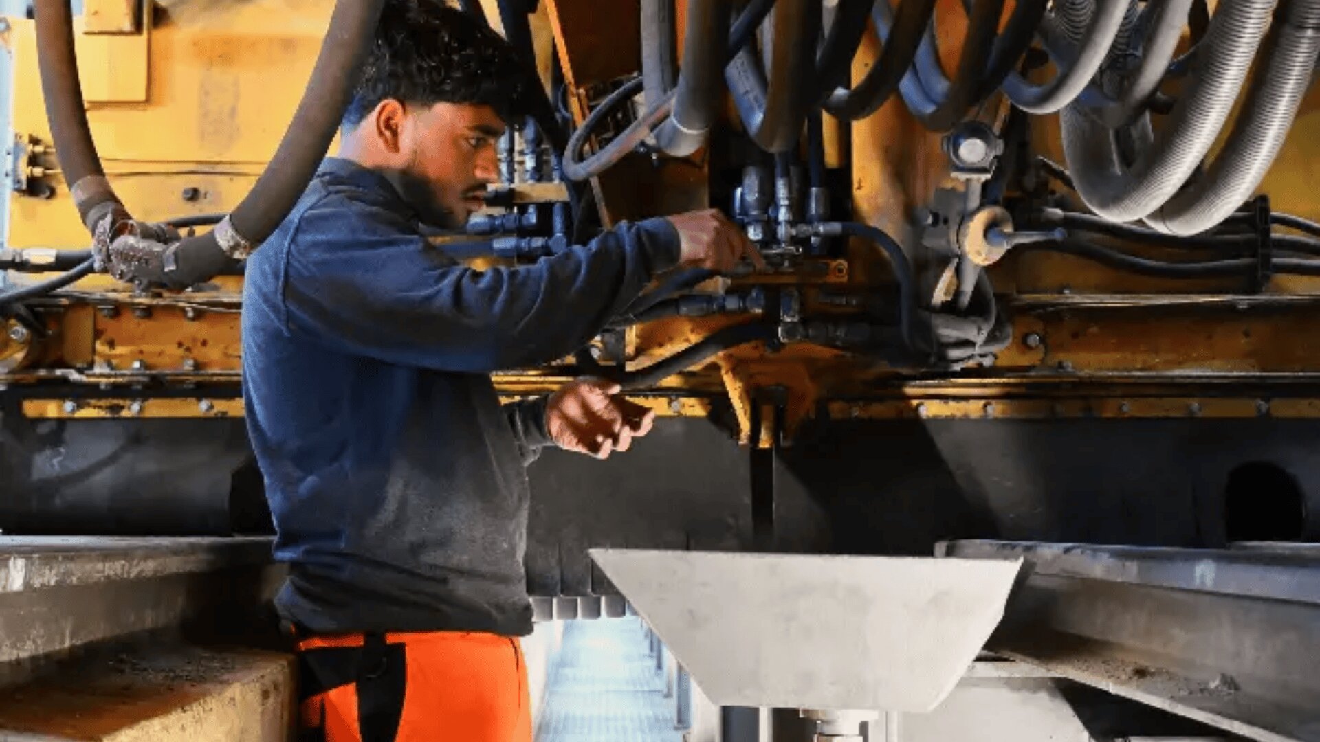 Ein Mann in einem dunklen Hemd und einer orangefarbenen Hose stellt Maschinen ein und prüft Rohre und Ventile in einer industriellen Umgebung.