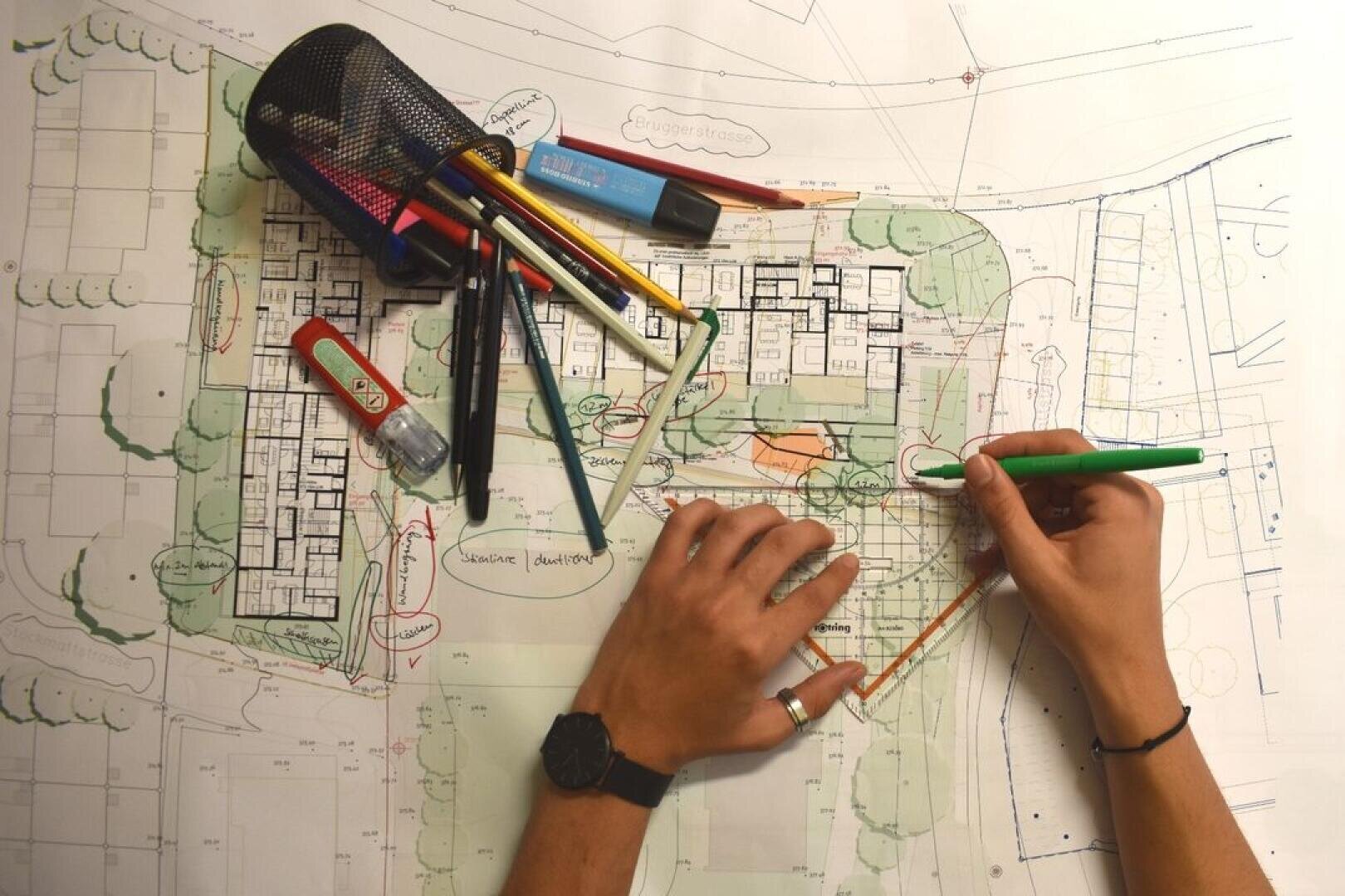Die Hände einer Person zeichnen mit einem grünen Stift und einem dreieckigen Lineal Linien auf Architekturpläne. Verschiedene farbige Stifte, Textmarker und Schreibwaren liegen auf dem Bauplan verstreut. Der Arbeitsbereich ist gut beleuchtet.