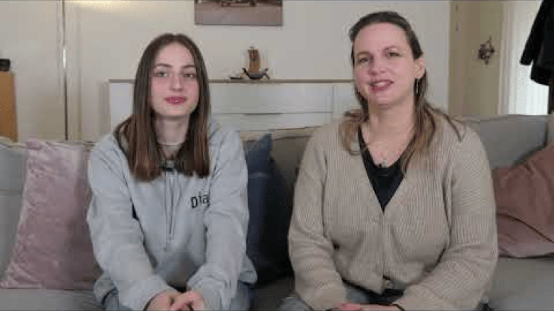 Zwei Frauen sitzen Seite an Seite auf einem hellen Sofa in einem Wohnzimmer. Eine trägt einen grauen Kapuzenpulli, die andere eine beigefarbene Strickjacke. Beide schauen in die Kamera und lächeln. Im Hintergrund sind Kissen und eine Kommode zu sehen.