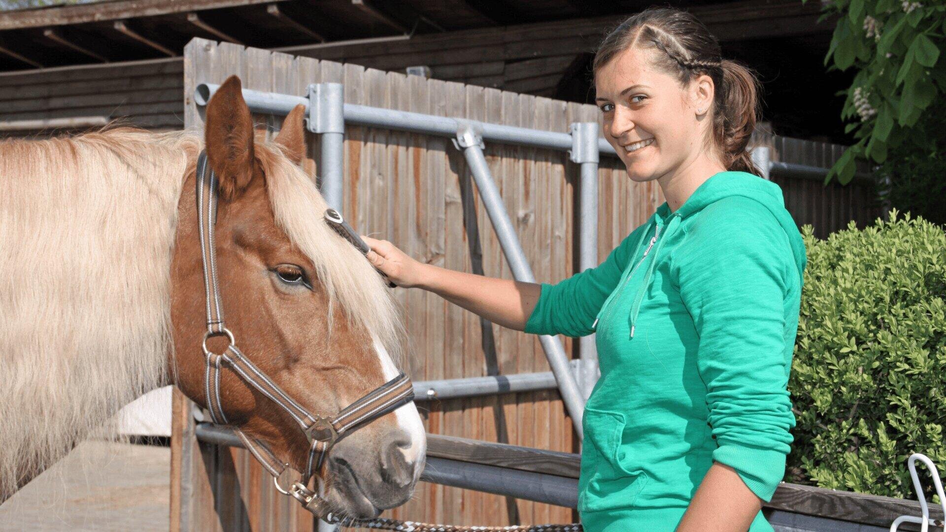 Eine Frau in einem grünen Kapuzenpulli lächelt und streichelt ein braunes Pferd mit weißer Mähne, das draußen an einem Holzzaun steht.