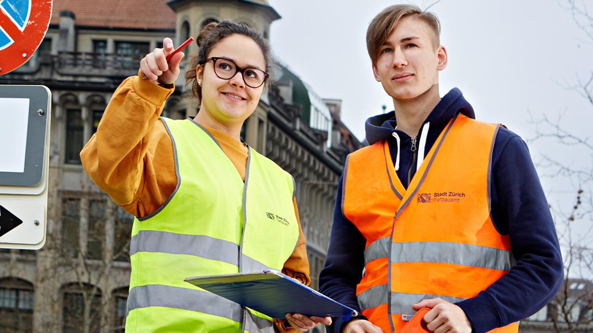 Zwei junge Leute mit reflektierenden Westen stehen im Freien; einer hält ein Klemmbrett und zeigt auf etwas, während der andere nach vorne schaut. Im Hintergrund sind Gebäude und Verkehrsschilder zu sehen.