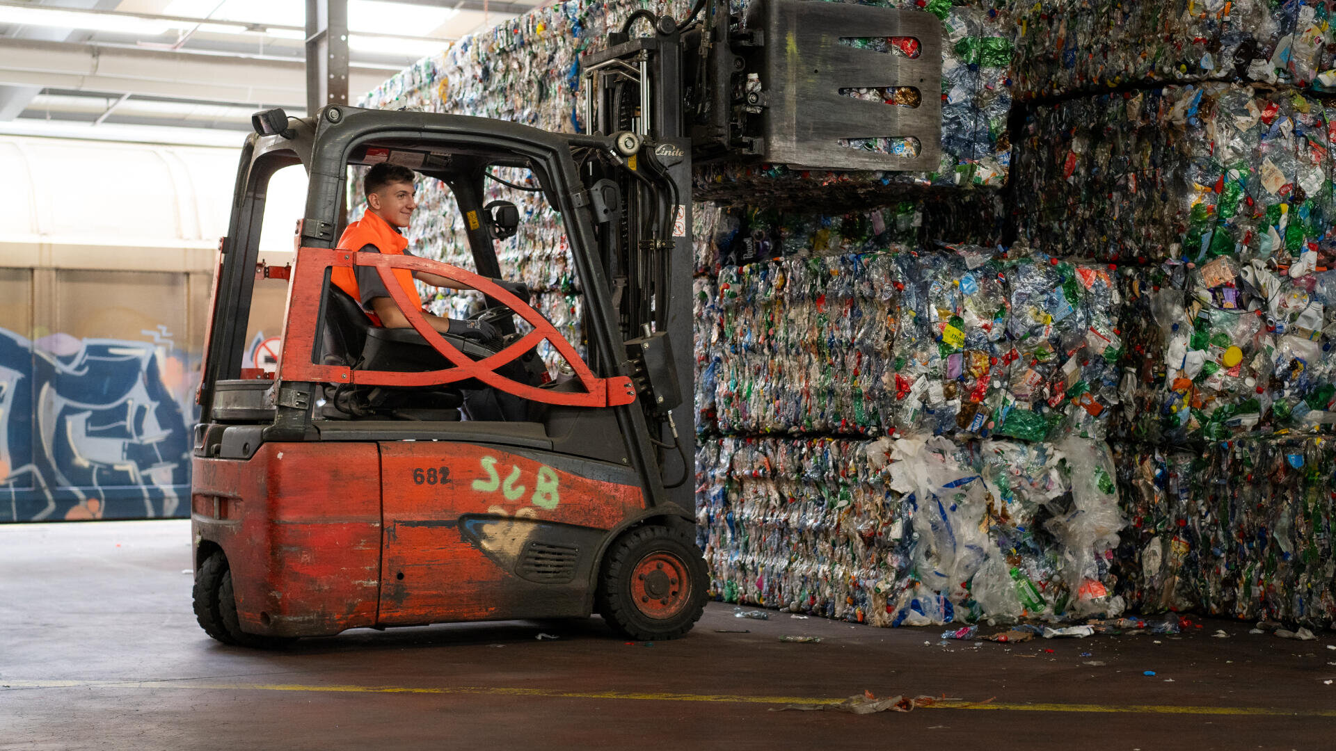 Ein Arbeiter in einem orangefarbenen Hemd bedient einen roten Gabelstapler und hebt große Ballen gepresster Plastikflaschen in einer Recyclinganlage. Im Hintergrund sind Stapel von Plastikballen und Graffiti zu sehen.
