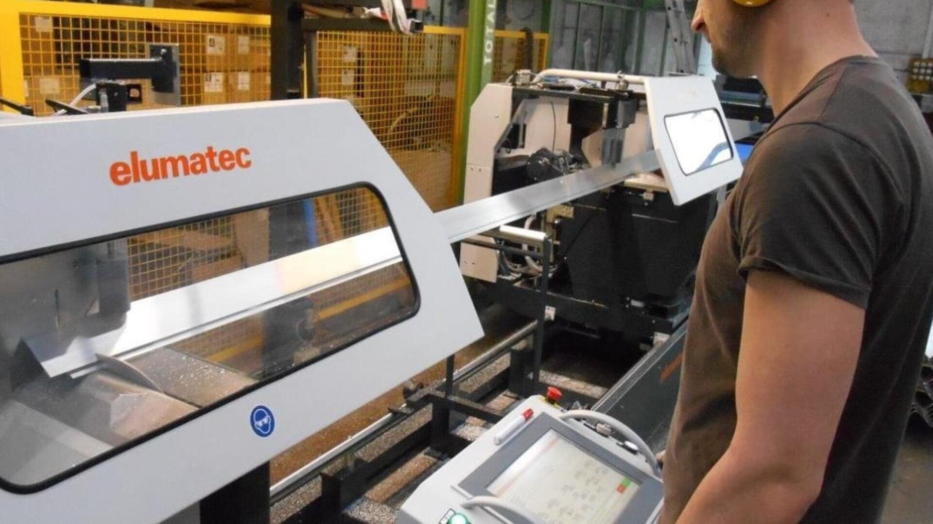 Eine Person, die einen Gehörschutz trägt, bedient eine Elumatec-Industriemaschine und überwacht das Bedienfeld, während ein langes Metallstück in einer Fabrikhalle bearbeitet wird.