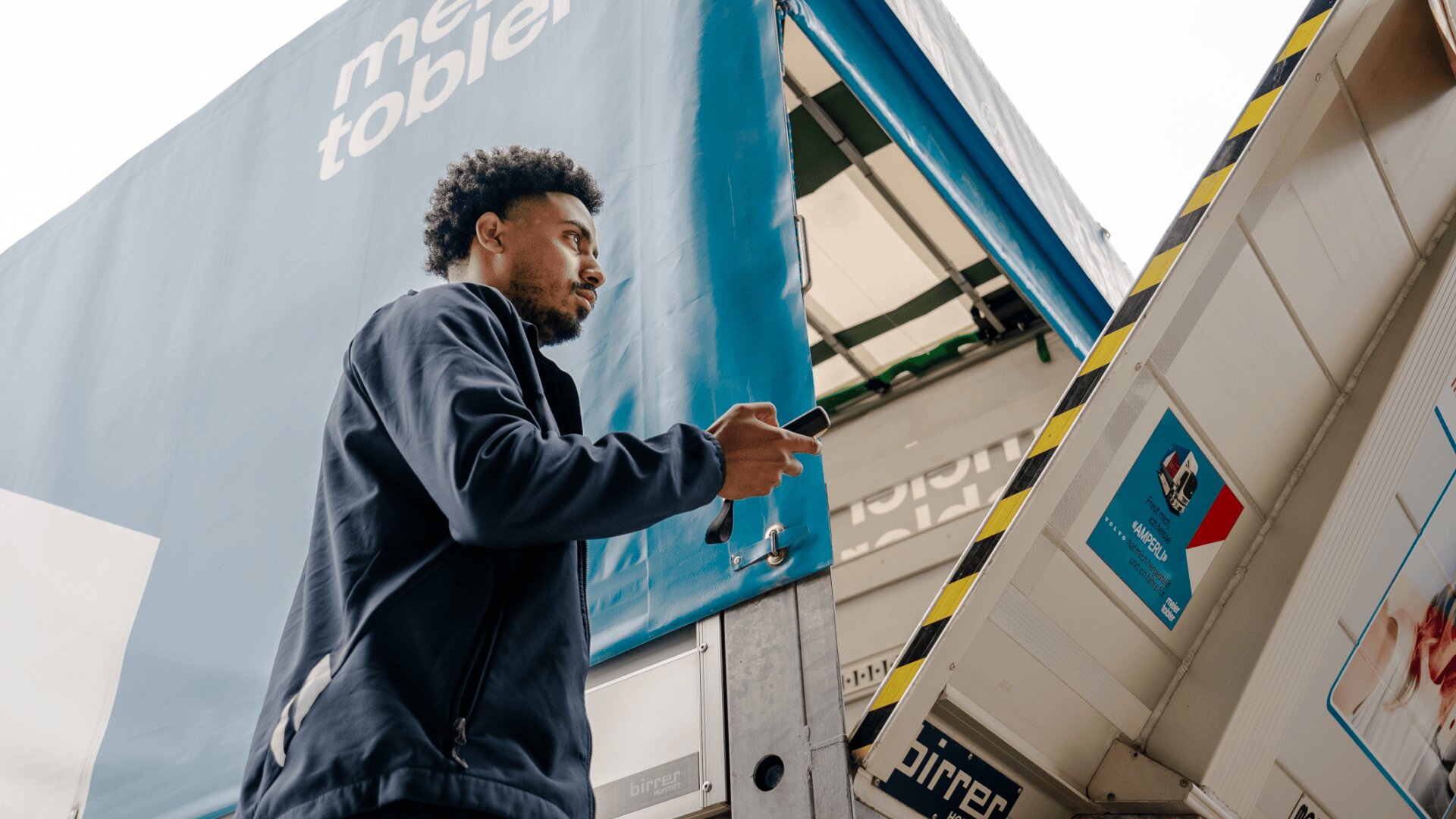 Ein Mann, der ein Smartphone in der Hand hält, steht neben einem offenen Lieferwagen und schaut nach oben, während er sich darauf vorbereitet, Pakete aus- oder einzuladen. Die Seite des blauen Lkw ist teilweise im Hintergrund zu sehen.