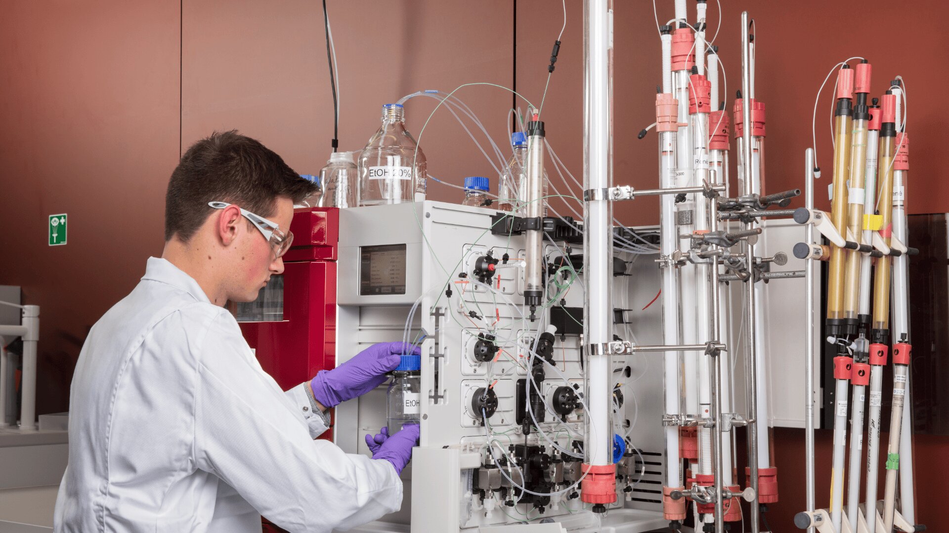 Ein Wissenschaftler mit weißem Laborkittel, Schutzbrille und violetten Handschuhen bedient eine komplexe Laborausrüstung mit Schläuchen und Drähten vor einer rötlich-braunen Wand.
