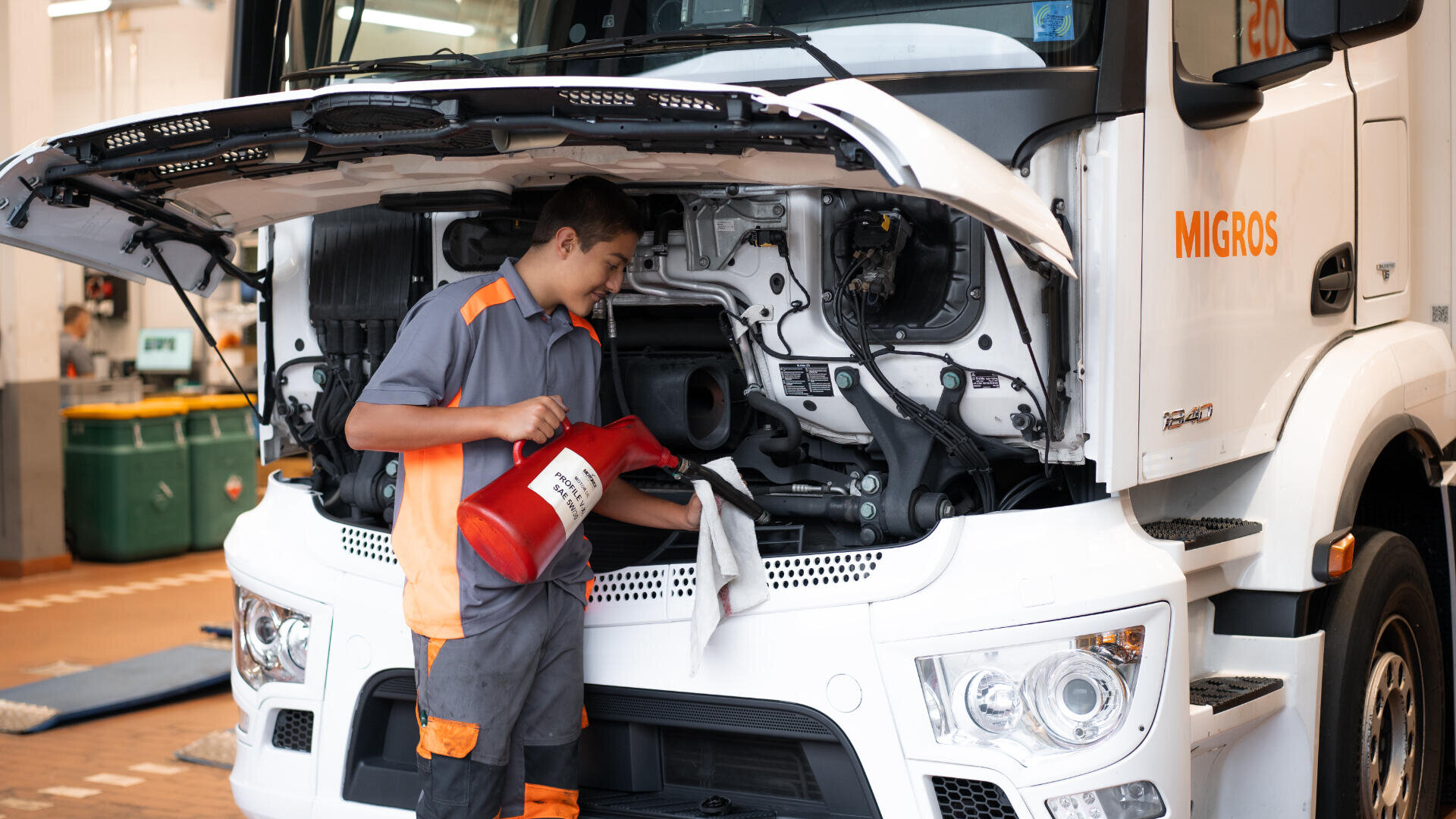 Ein Mechaniker in grauer und orangefarbener Uniform gießt bei geöffneter Motorhaube Öl in einen Lkw-Motor, während er einen roten Ölbehälter und ein weißes Tuch hält. Auf der Seite des Lastwagens steht "Migros".