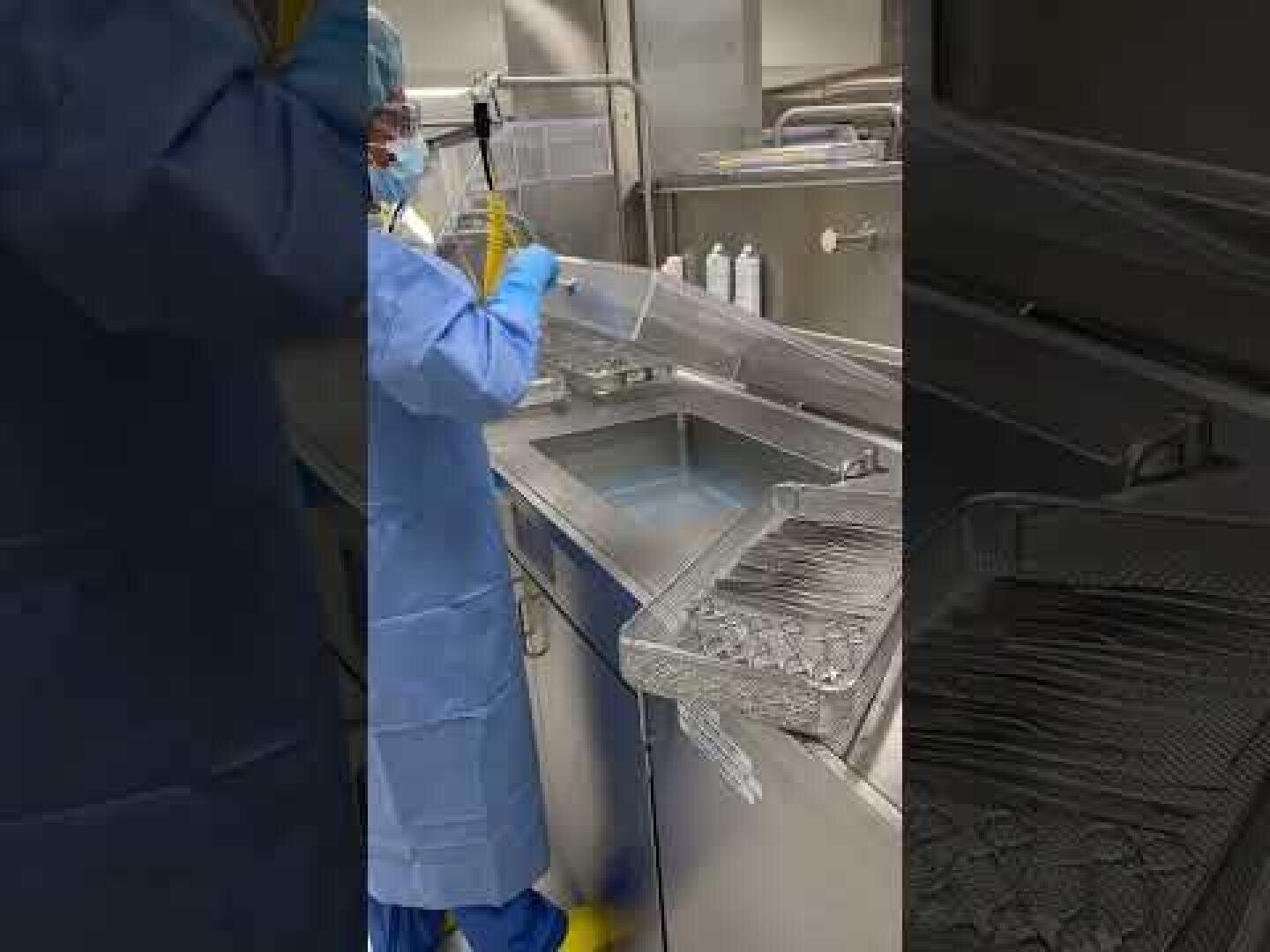 Eine Person in Schutzkleidung und mit Handschuhen reinigt mit einem Sprühschlauch medizinische Instrumente an einem Waschbecken aus Edelstahl in einer Klinik oder einem Labor.