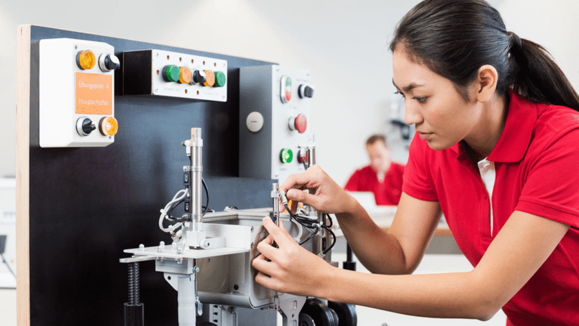 Eine junge Frau in einem roten Polohemd justiert ein Bauteil an einer Industriemaschine in einer Laborumgebung und konzentriert sich dabei auf ihre Arbeit. Im Hintergrund sind Schalttafeln mit Tasten und Schaltern zu sehen.