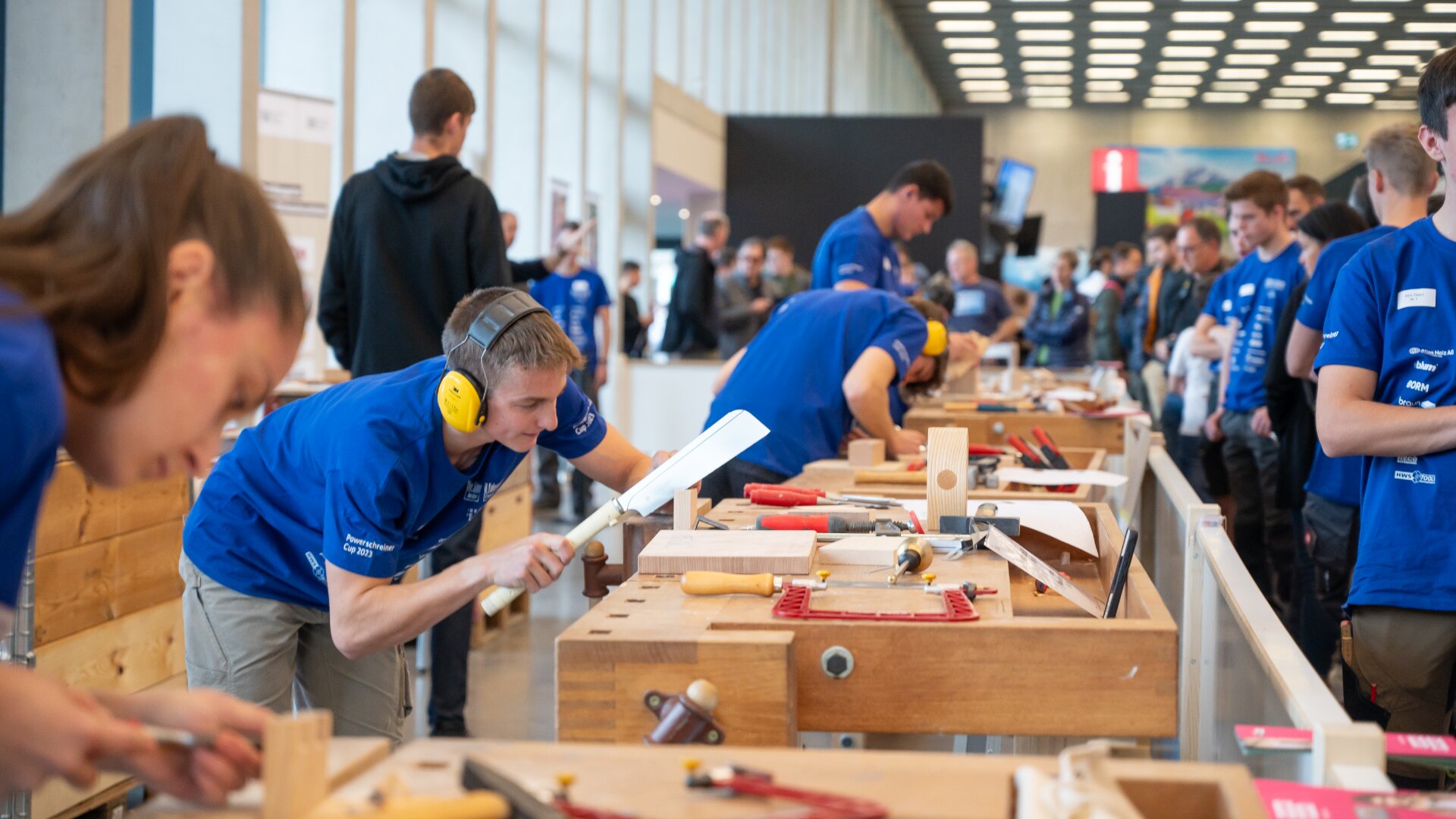 Junge Leute in blauen Hemden nehmen an einem Holzbearbeitungswettbewerb teil und arbeiten an Bänken mit Werkzeugen. Eine Person im Vordergrund bearbeitet Holz mit einem Hobel und trägt dabei einen Gehörschutz. Im Hintergrund schauen viele Menschen zu.