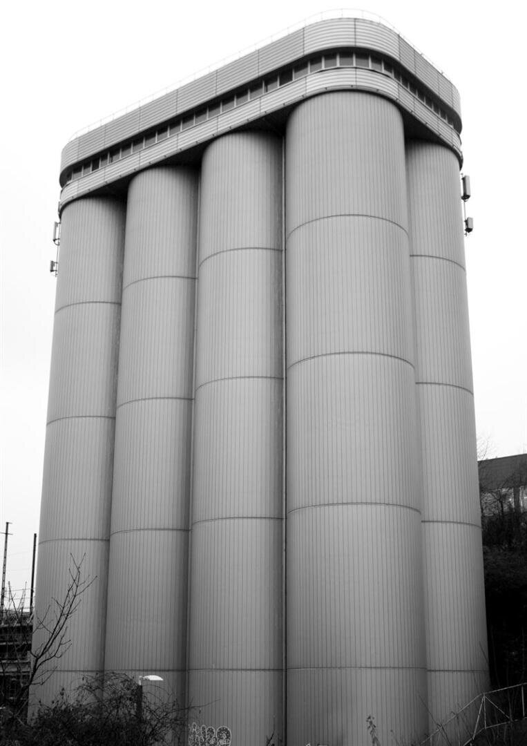 Ein Schwarz-Weiß-Foto eines hohen Industriegebäudes mit sechs großen, vertikalen zylindrischen Silos und einem abgerundeten Dach, umgeben von spärlicher Vegetation und einem bewölkten Himmel.
