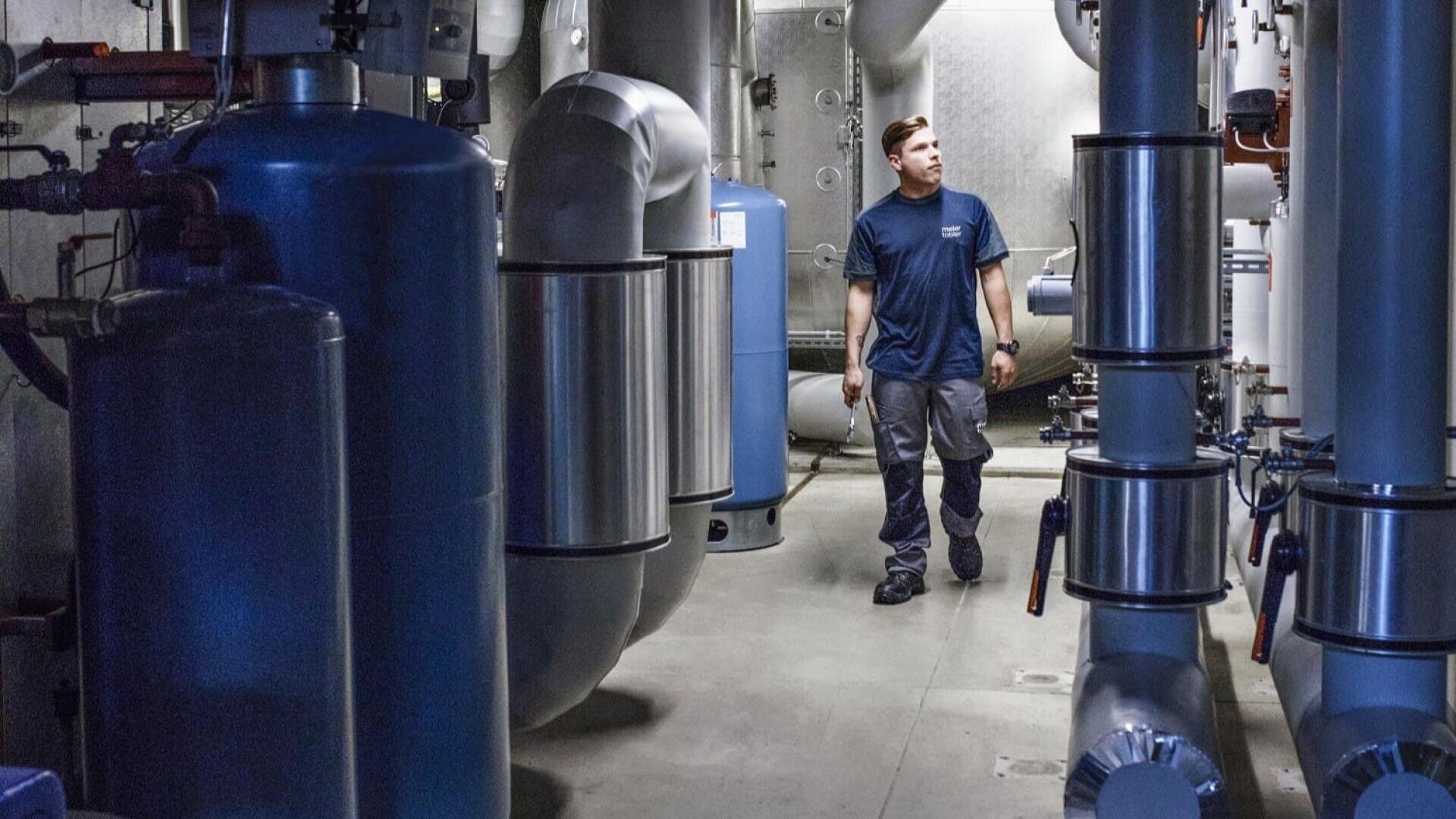 Ein Mann in blauem Hemd und grauer Hose geht durch einen Raum voller großer Industrierohre, Tanks und Maschinen und inspiziert die Ausrüstung in einem Versorgungs- oder Maschinenraum.