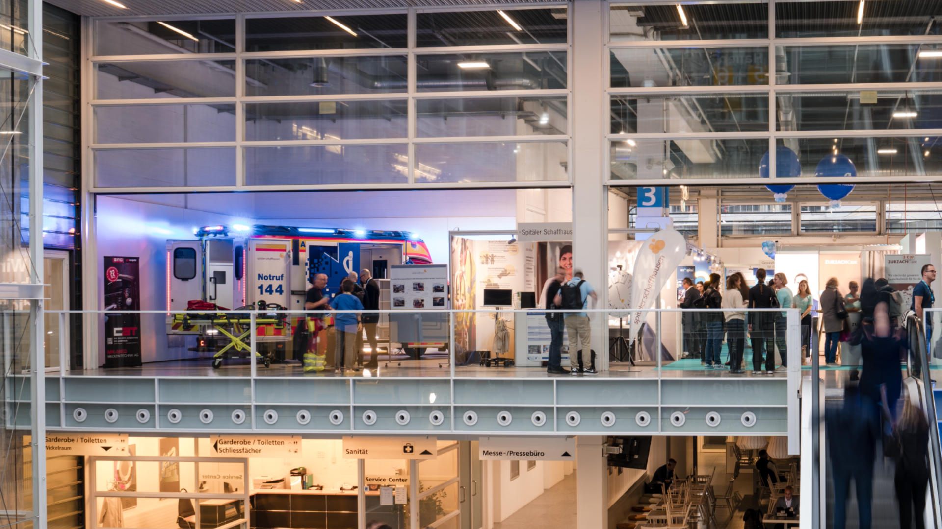 Eine moderne Ausstellungshalle mit Glaswänden, mehreren Ebenen und Menschen, die herumlaufen und sich versammeln. Ein Krankenwagen mit Blinklichtern steht im Obergeschoss inmitten verschiedener Stände und Informationsdisplays.