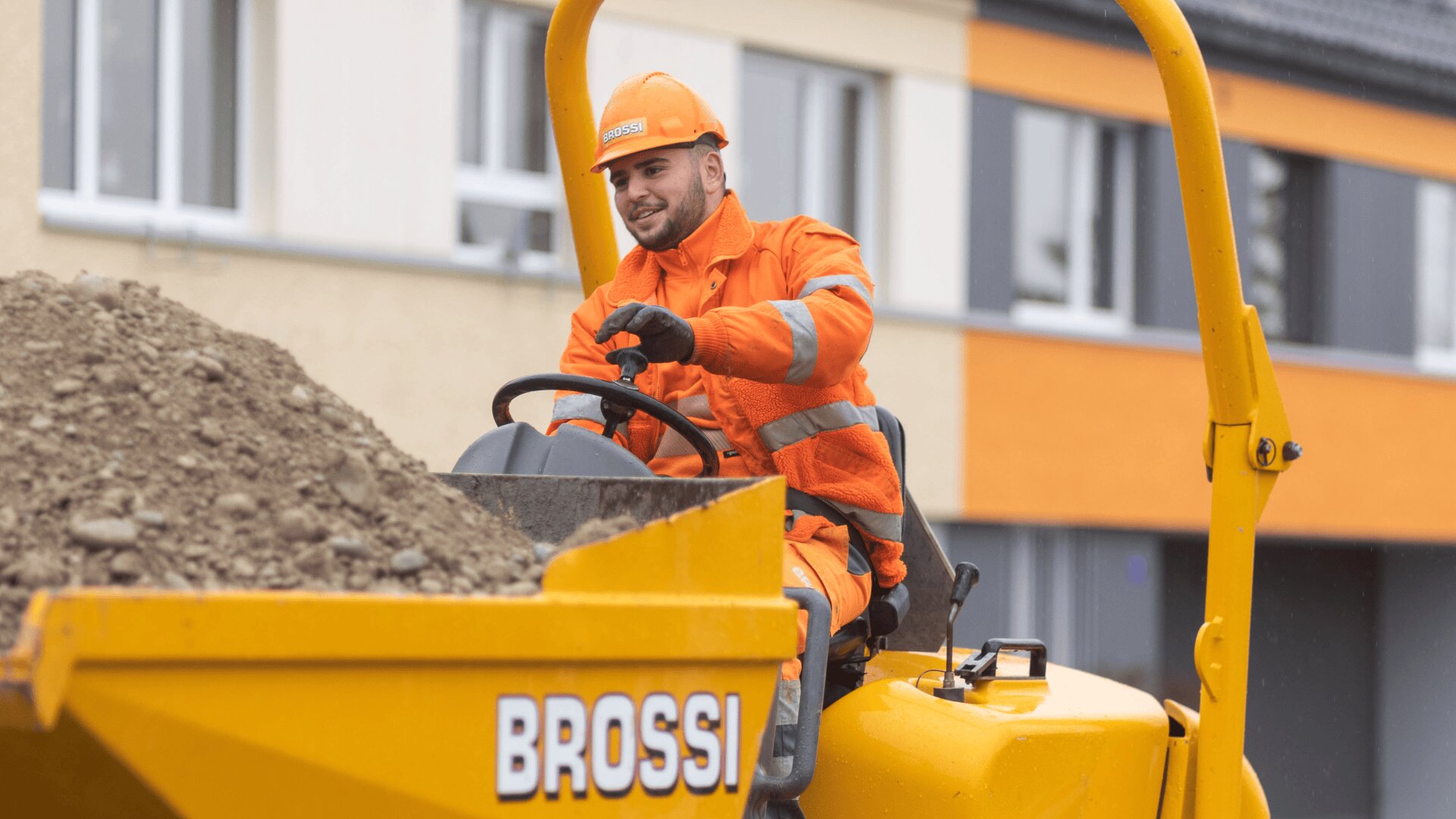 Ein Bauarbeiter in orangefarbener Schutzkleidung und Helm bedient einen gelben Brossi-Muldenkipper, der mit Erde gefüllt ist. Er lächelt, im Hintergrund sind ein Gebäude und Fenster zu sehen.