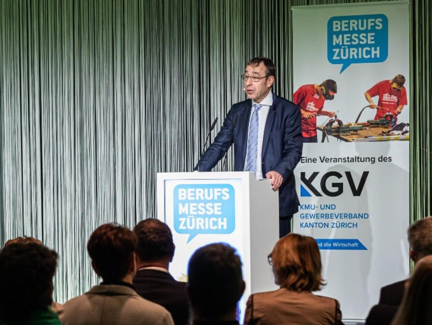 Ein Mann im Anzug spricht auf einem Podium mit der Aufschrift Berufs Messe Zürich vor einem Publikum, im Hintergrund ein Banner für die Berufs Messe Zürich und KGV.