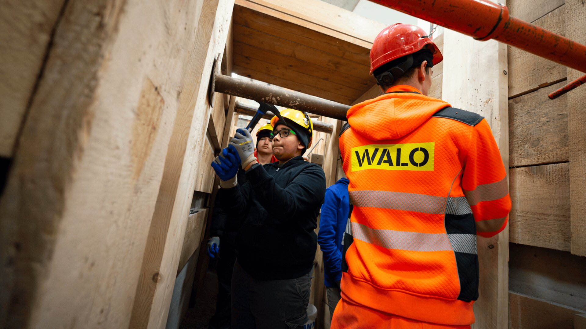 Mehrere Bauarbeiter, die Schutzkleidung und Helme tragen, arbeiten in einem schmalen, holzverkleideten Graben. Ein Arbeiter in einer orangefarbenen Jacke mit der Aufschrift WALO steht mit dem Rücken zur Kamera im Vordergrund.