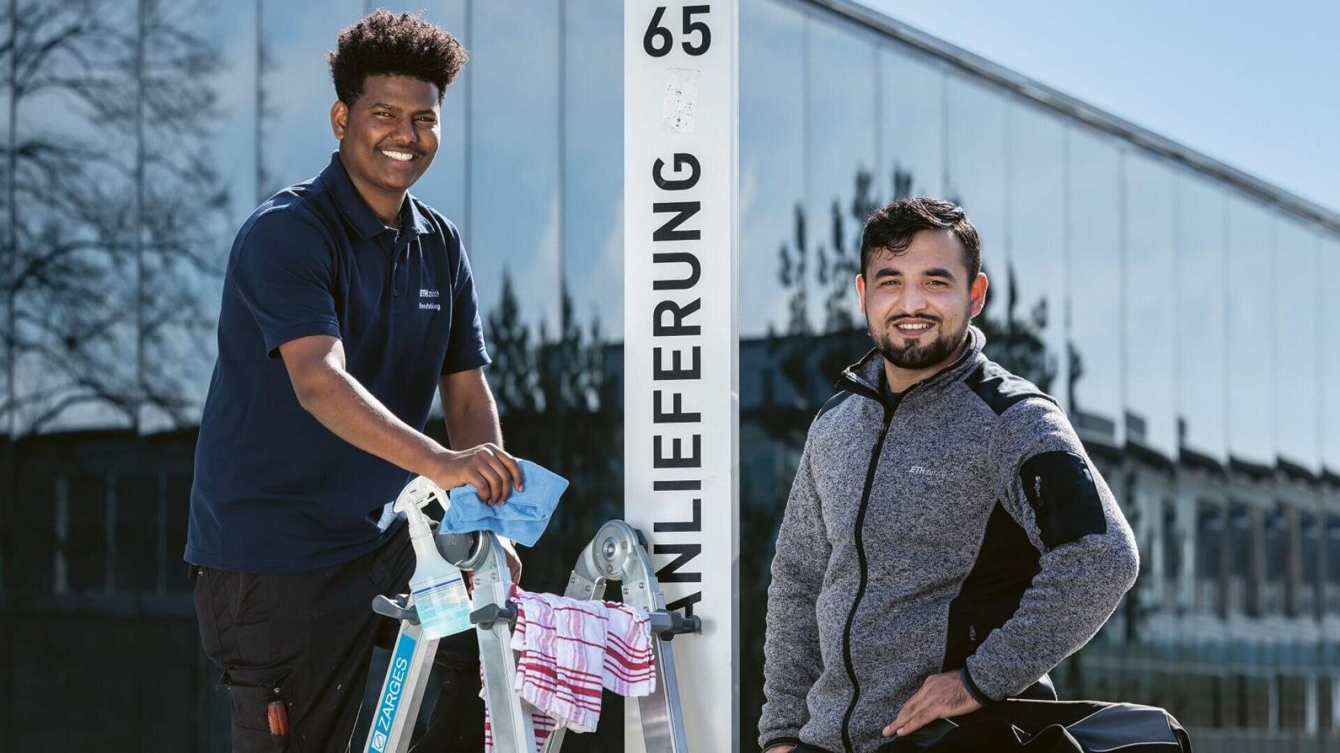 Zwei Männer lächeln im Freien neben einem Schild mit der Aufschrift "Anlieferung 65". Ein Mann steht mit einer Leiter und Reinigungsmitteln, während der andere daneben steht. Im Hintergrund ist ein modernes Glasgebäude zu sehen.