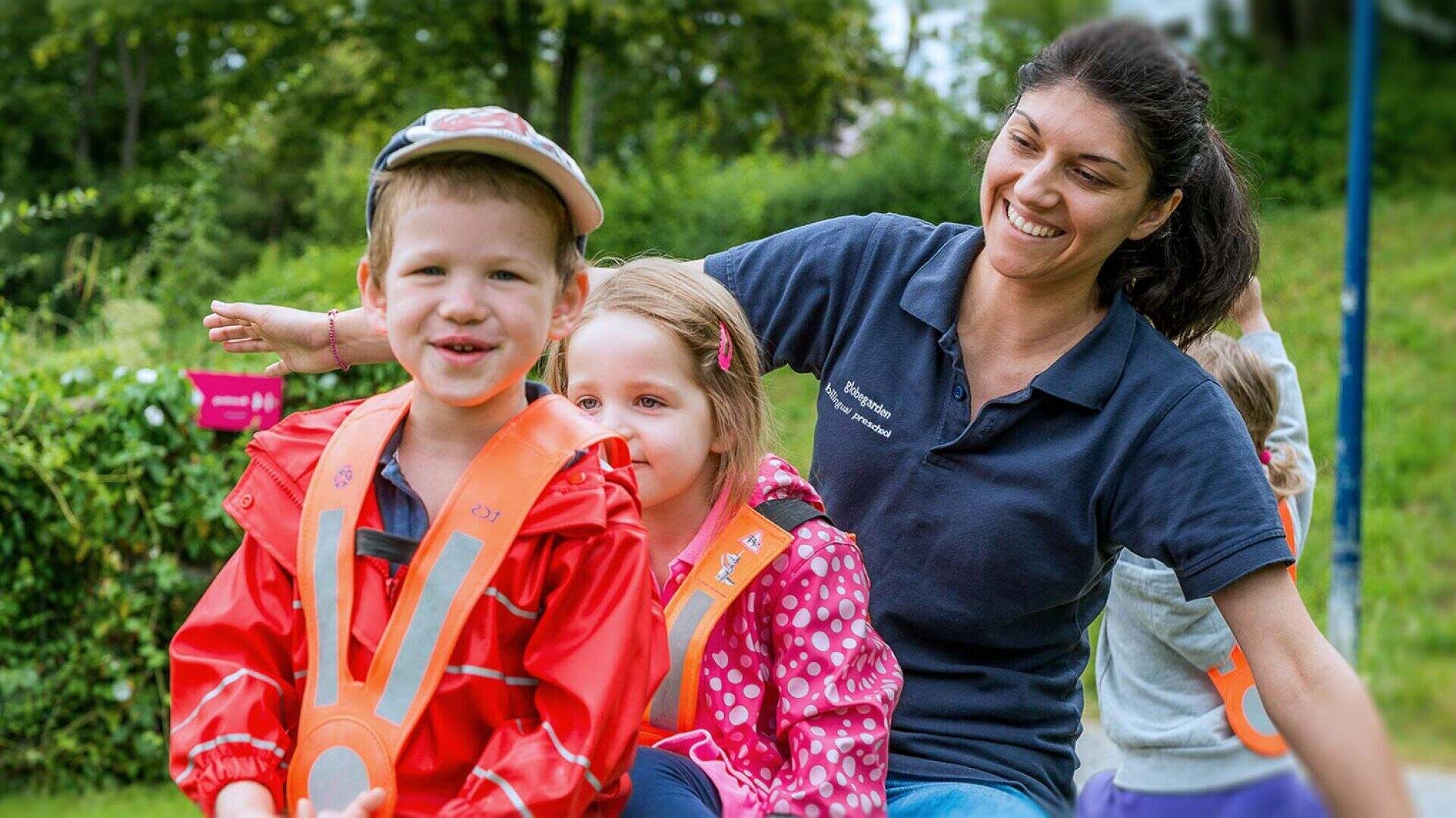 Eine lächelnde Frau sitzt im Freien mit einer Gruppe kleiner Kinder. Zwei Kinder im Vordergrund tragen bunte Regenjacken und orangefarbene Sicherheitswesten. Die Gruppe scheint eine spielerische Aktivität in einer grünen, parkähnlichen Umgebung zu genießen.