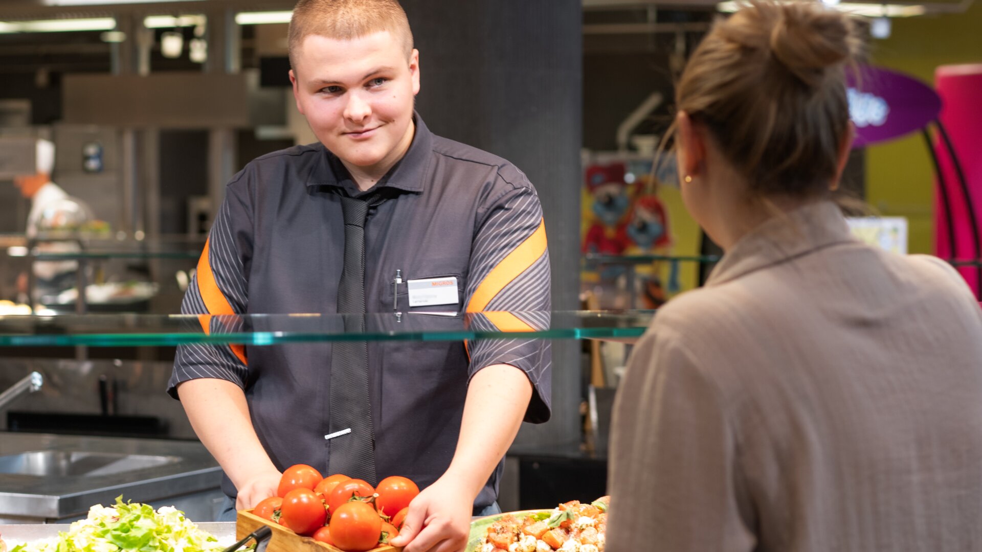 Ein Mann in schwarzer Uniform serviert einer Frau an einer Cafeteria-Theke frische Tomaten. Auf dem Tresen stehen verschiedene Lebensmittel, und im Hintergrund ist ein hell erleuchteter Essbereich zu sehen.