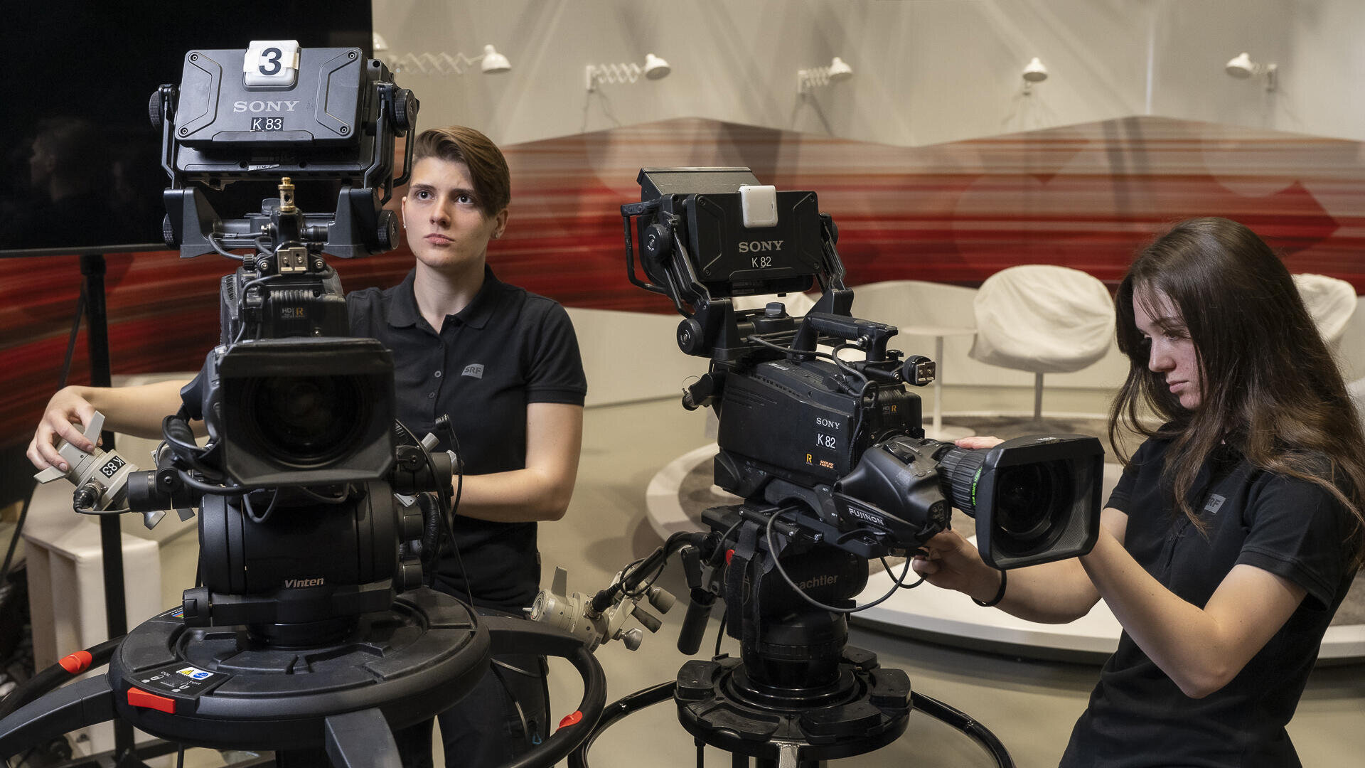 Zwei Frauen bedienen professionelle Videokameras in einem Studio mit weißen Stühlen und einer rot gestreiften Wand im Hintergrund. Beide sind mit der Einstellung der Ausrüstung beschäftigt und tragen schwarze Hemden.