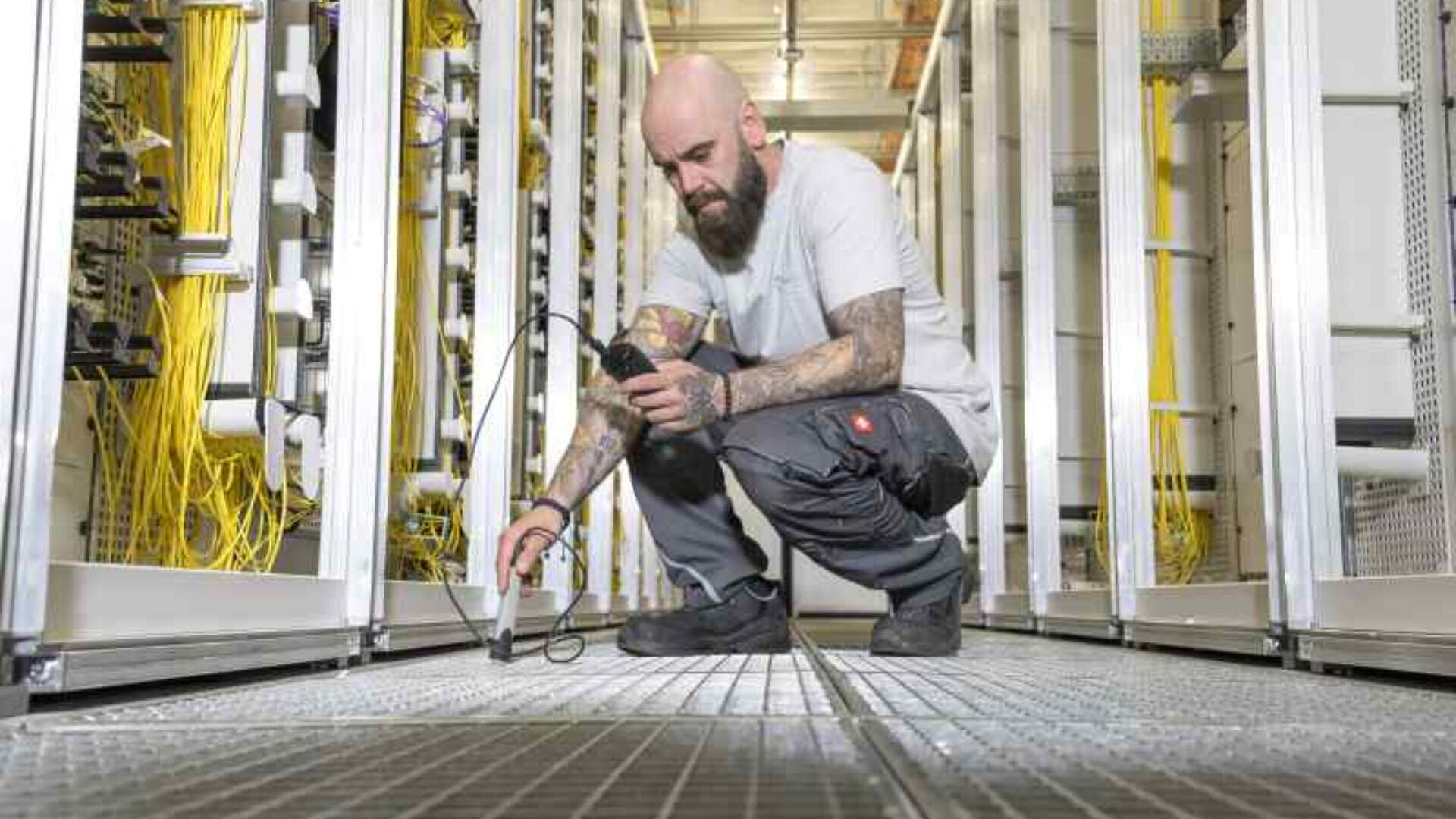Ein Techniker mit Tattoos und Bart hockt in einem Serverraum und überprüft mit einem Handheld-Gerät die Geräte. Gelbe Kabel säumen beide Seiten des Ganges, und der Metallboden ist sichtbar.