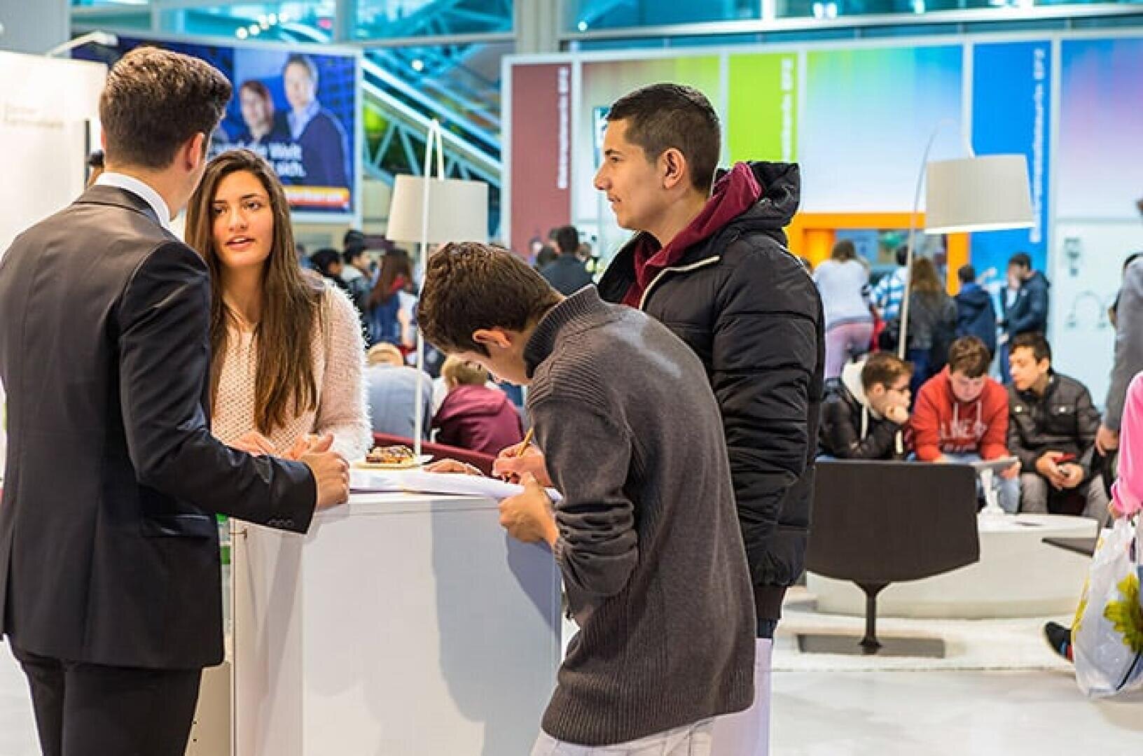 Eine Gruppe junger Erwachsener unterhält sich an einem weißen Stand bei einer belebten Veranstaltung in einer Halle. Im Hintergrund sitzen und gehen Menschen, und bunte Tafeln und Lichter schaffen eine lebendige Atmosphäre.