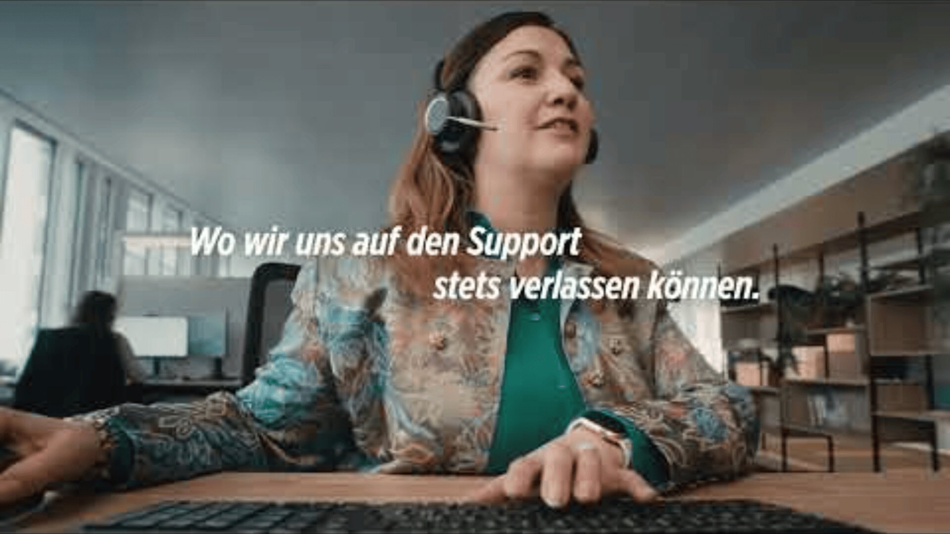 Eine Frau, die ein Headset trägt, sitzt an einem Schreibtisch und benutzt einen Computer in einem modernen Büro. Der Text auf Deutsch über dem Bild lautet: "Wo wir uns auf den Support stets verlassen können.