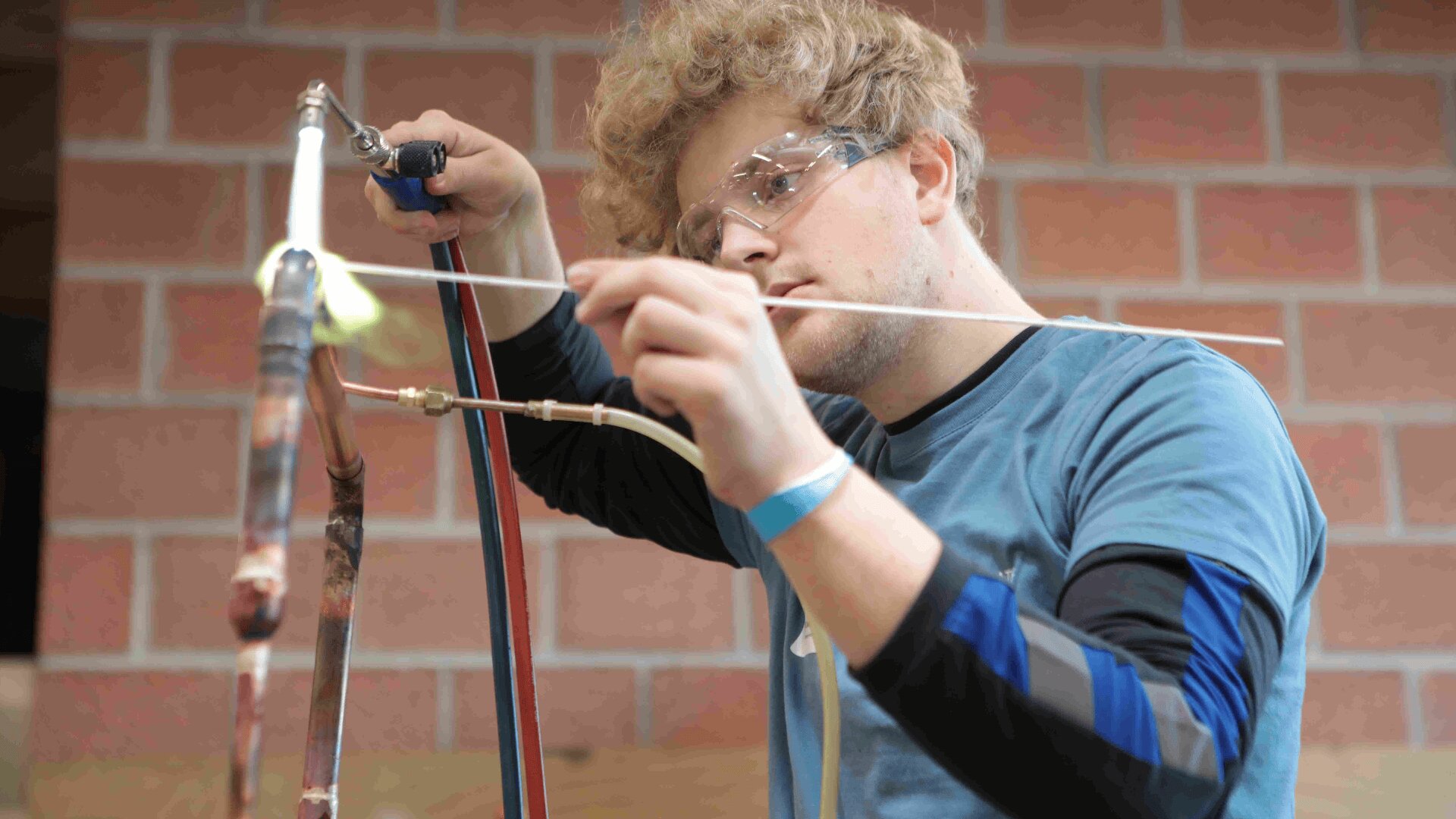 Ein junger Mann mit lockigem Haar und Schutzbrille arbeitet sorgfältig und konzentriert mit Metallrohren und Werkzeugen. Er befindet sich in einem Innenraum, mit einer Ziegelmauer im Hintergrund.