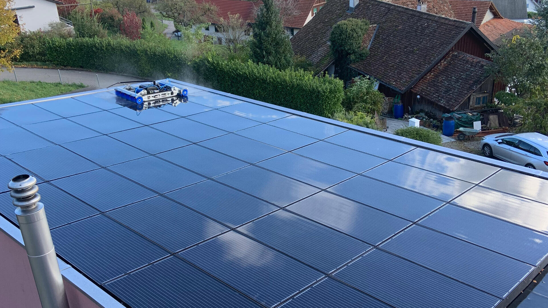 Ein mit Solarpaneelen bedecktes Dach wird von einem kleinen, blauen automatischen Reinigungsroboter gereinigt. Im Hintergrund sind Häuser, Gärten und geparkte Autos zu sehen.