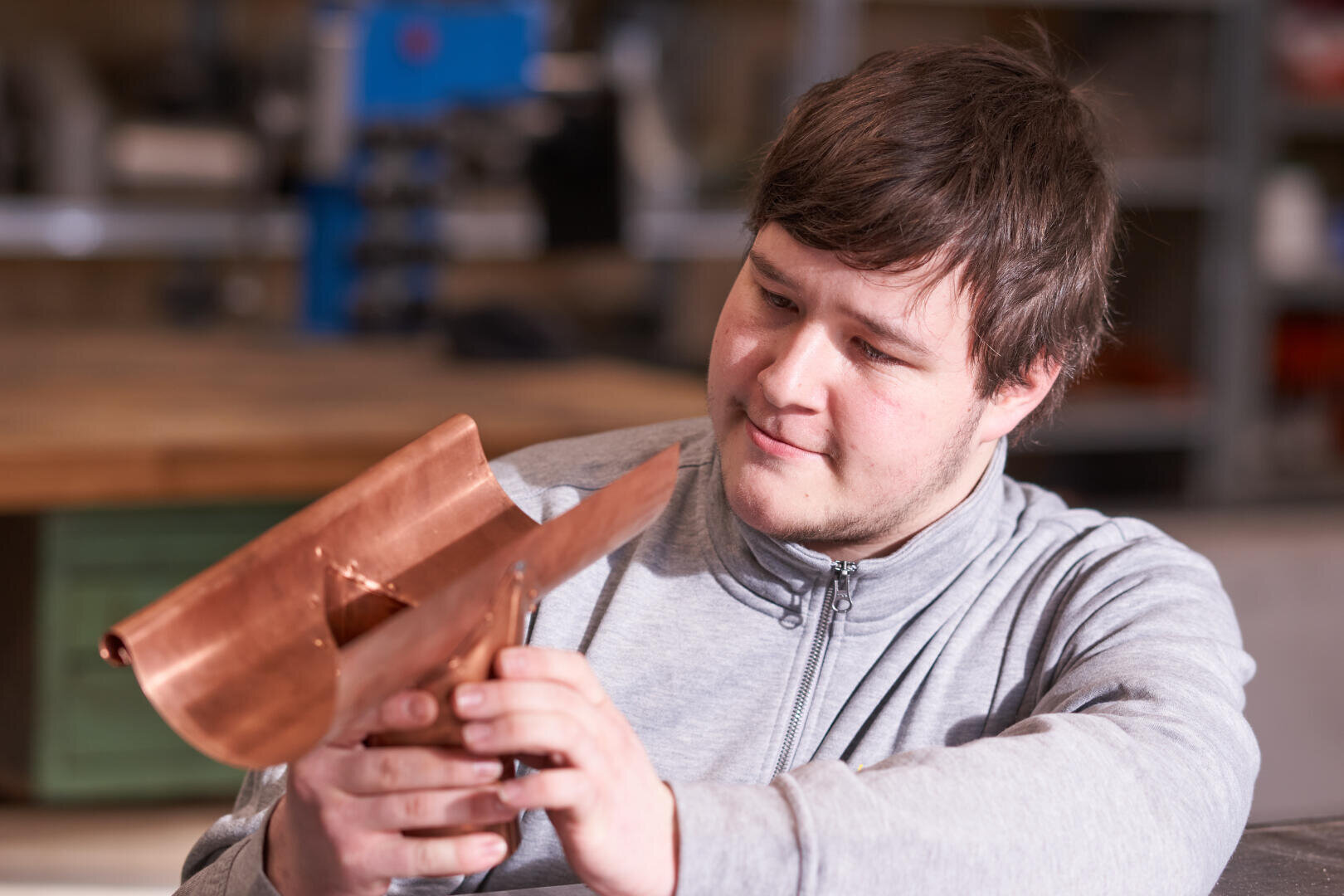 Eine Person in einer grauen Jacke mit Reißverschluss untersucht in einer Werkstattumgebung mit Regalen und Werkzeugen im Hintergrund einen Metallgegenstand, möglicherweise ein handgefertigtes Bauteil oder eine Skulptur.