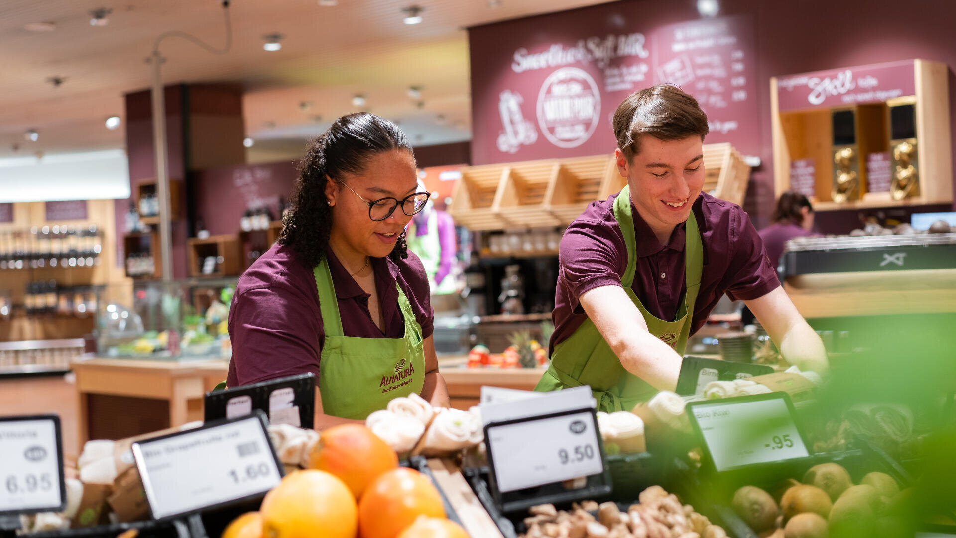 Zwei Angestellte eines Lebensmittelgeschäfts mit grünen Schürzen und violetten Hemden sortieren in einem gut beleuchteten Supermarkt frische Produkte, darunter Pilze und Orangen, in der Gemüseabteilung.