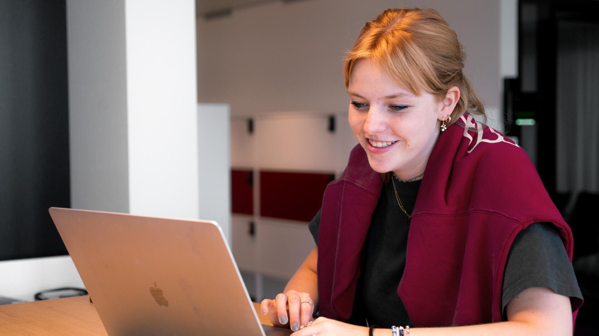 Eine junge Frau mit hellem Haar arbeitet lächelnd an einem Laptop an einem Schreibtisch. Sie trägt ein dunkles Hemd mit einem kastanienbraunen Pullover, der über ihre Schultern drapiert ist, in einem modernen Innenraum.