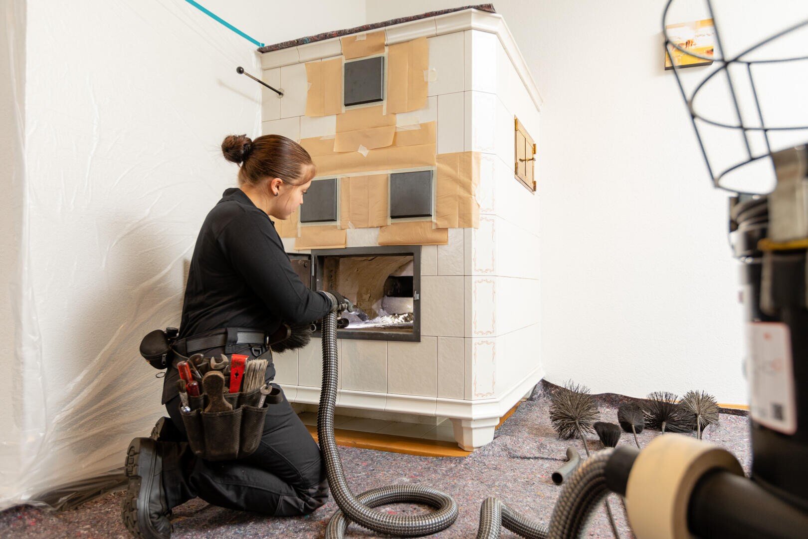Eine Person in schwarzer Arbeitskleidung kniet beim Reinigen eines Kachelkamins mit einem Staubsaugerschlauch. Die Werkzeuge befinden sich in ihrem Gürtel, und die Bürsten liegen neben ihr auf dem Boden. Der Kamin ist teilweise mit Abdeckband abgedeckt.