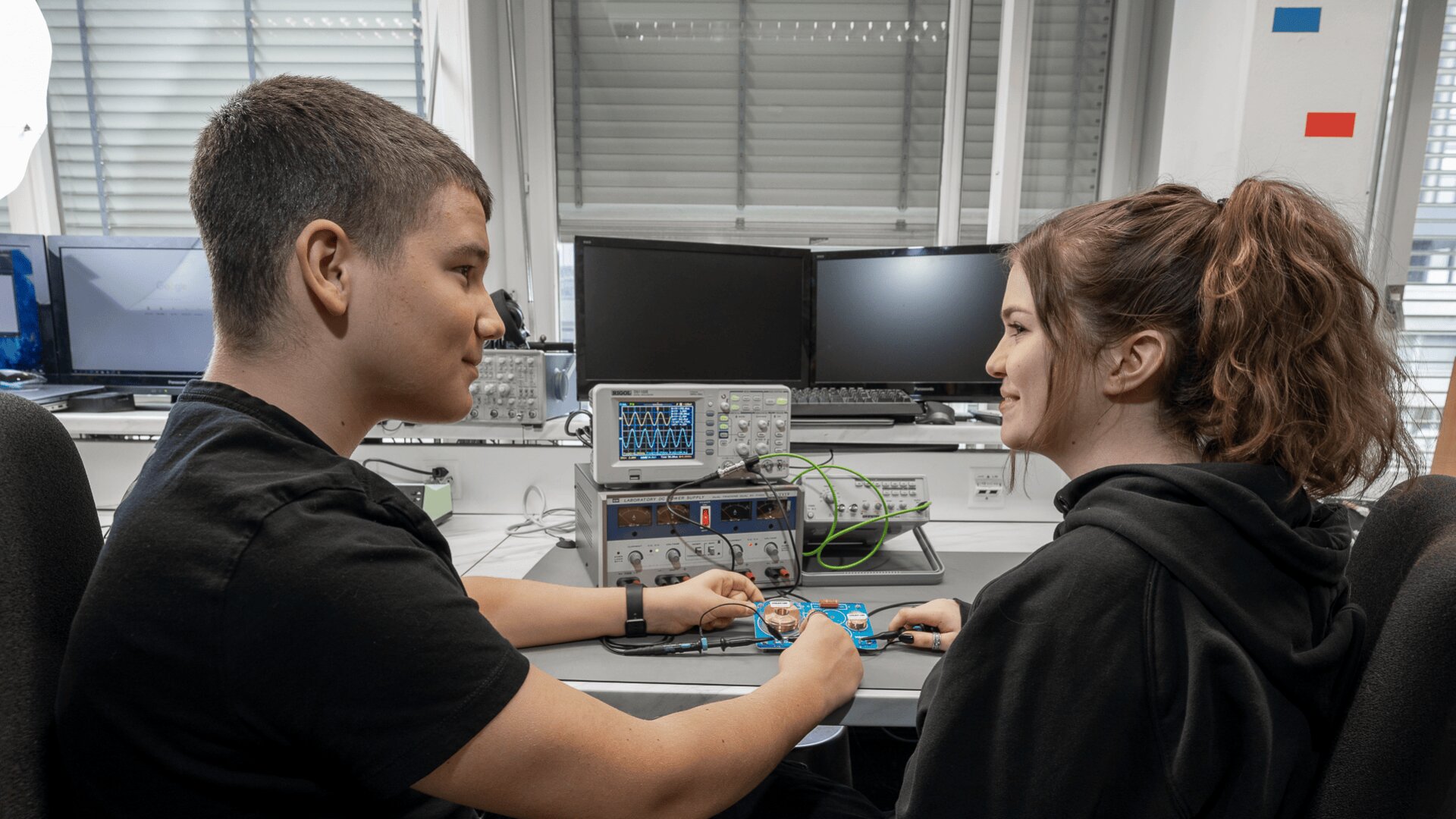 Zwei junge Menschen sitzen sich in einem Labor gegenüber und halten Sensoren, die mit elektronischen Geräten auf einem Tisch verbunden sind. Im Hintergrund sind Computerbildschirme und technische Geräte zu sehen.