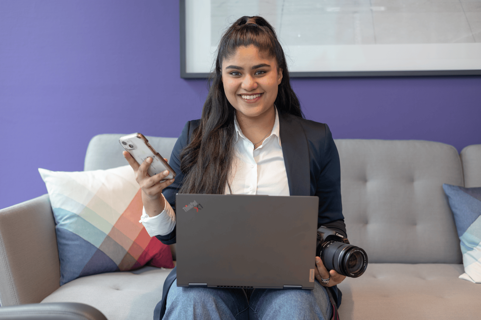 Eine Frau sitzt auf einem grauen Sofa, hält ein Telefon und eine Kamera in der Hand und hat einen Laptop auf dem Schoß. Sie lächelt, trägt einen Blazer und ein weißes Hemd, neben ihr ein buntes Kissen und im Hintergrund eine lila Wand.