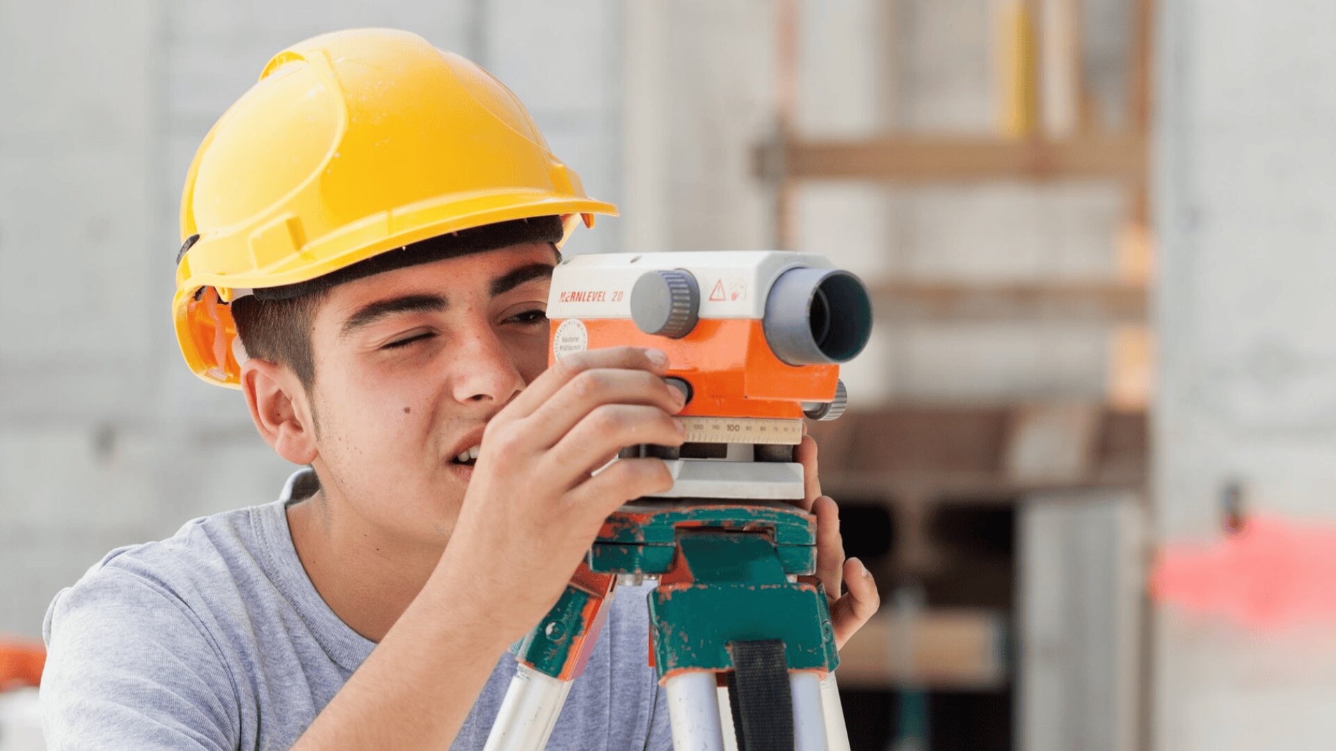 Ein junger Mann mit einem gelben Schutzhelm benutzt auf einer Baustelle ein Vermessungsinstrument auf einem Stativ und stellt das Gerät konzentriert ein.