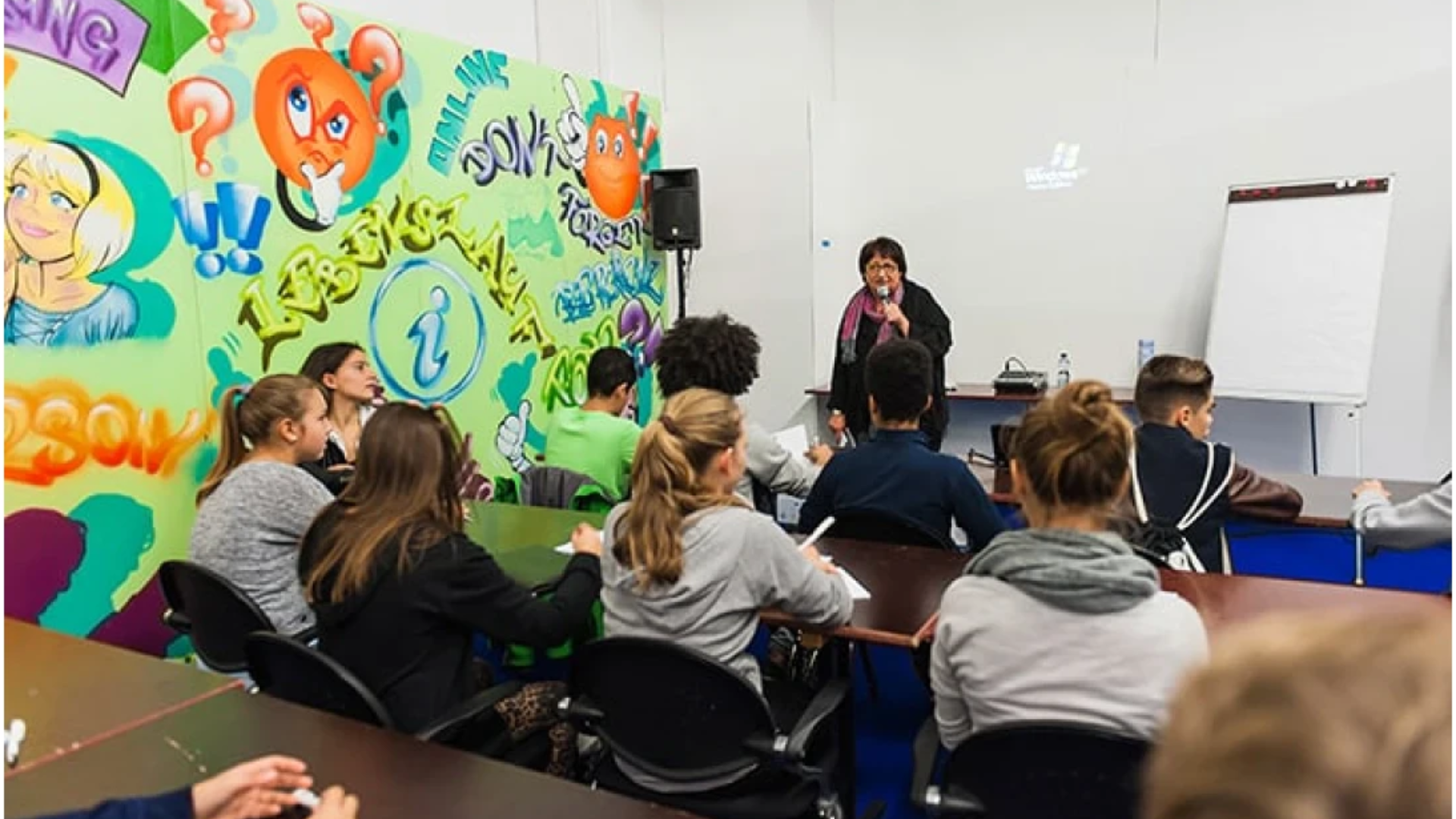 Ein Klassenzimmer mit Schülern, die an Tischen sitzen und einer Lehrerin gegenübersitzen, die an der Stirnseite spricht. An der Wand hinter ihr befinden sich bunte Graffiti-Kunstwerke mit Cartoon-Gesichtern und Fragezeichen.