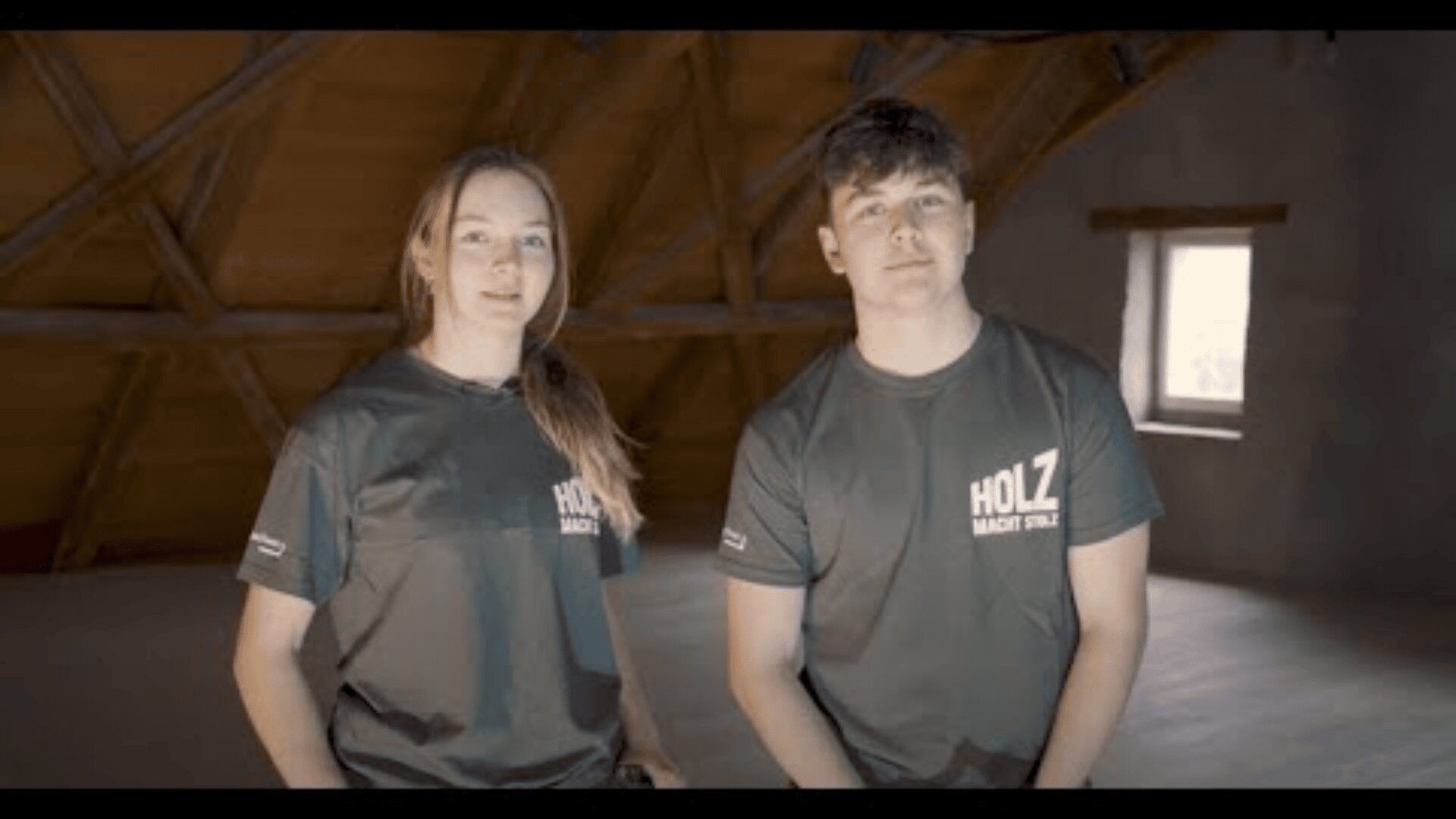 Zwei junge Leute in passenden dunklen T-Shirts mit dem Aufdruck HOLZ MACHT STOLZ stehen in einem geräumigen Dachgeschoss mit Holzbalken und einem einzigen Fenster.