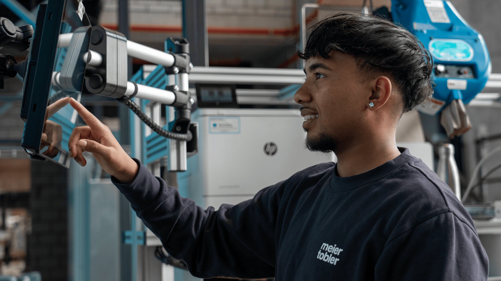 Eine Person in einem schwarzen Meijer-Tobler-Hemd bedient ein Touchscreen-Panel in einer industriellen Umgebung mit Maschinen im Hintergrund.