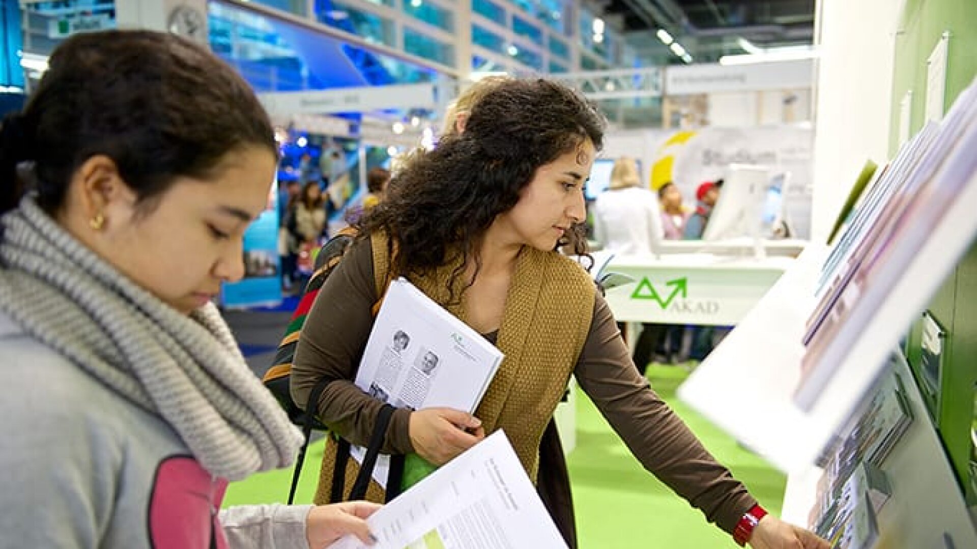 Zwei Frauen betrachten Broschüren und Papiere an einem Stand während einer Indoor-Veranstaltung oder Messe. Eine Frau hält Dokumente und greift nach weiteren Materialien, während die andere Frau in der Nähe steht und liest. Im Hintergrund sind andere Teilnehmer und Auslagen zu sehen.