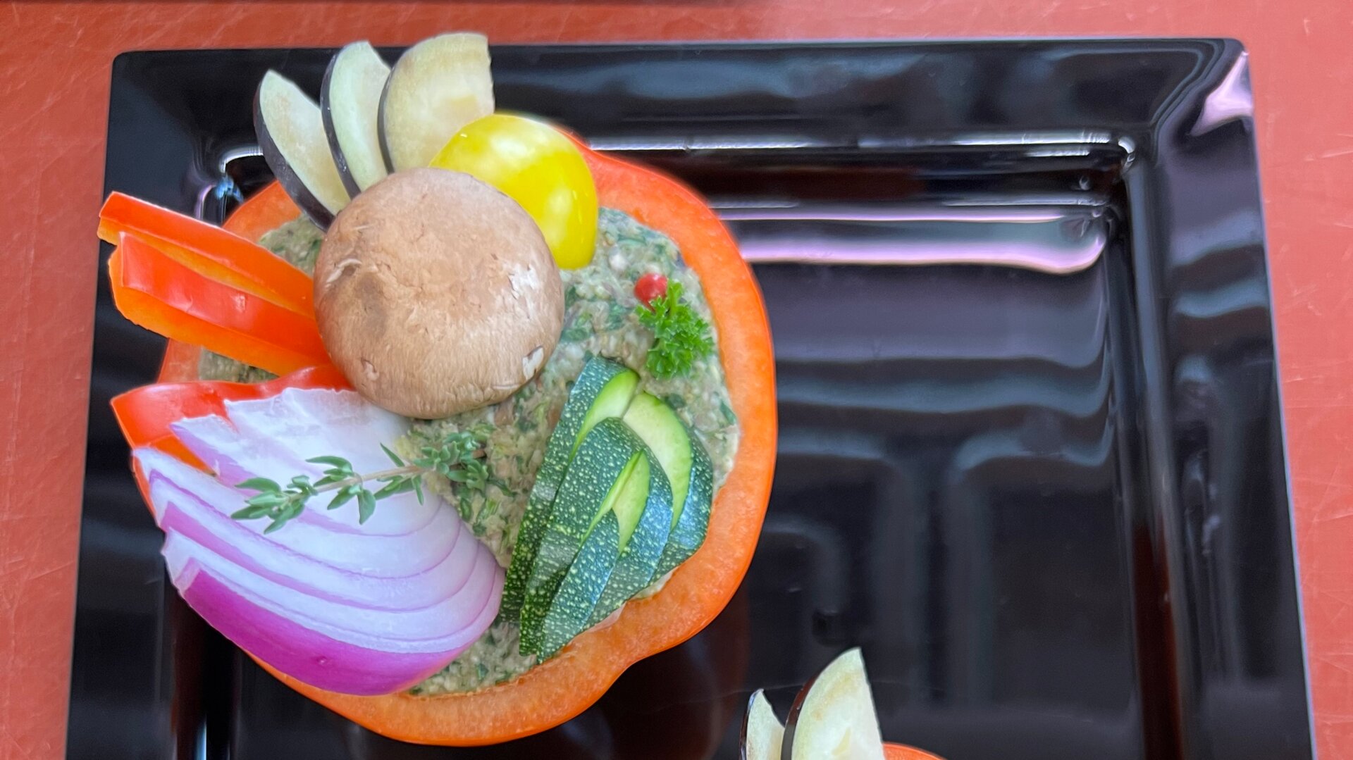Eine rote Paprikahälfte, gefüllt mit einem cremigen Dip, belegt mit geschnittenen Zucchini, roten Zwiebeln, roten Paprikastreifen, einem ganzen Champignon, einer gelben Kirschtomate und Kräutern, serviert auf einem schwarzen Teller.