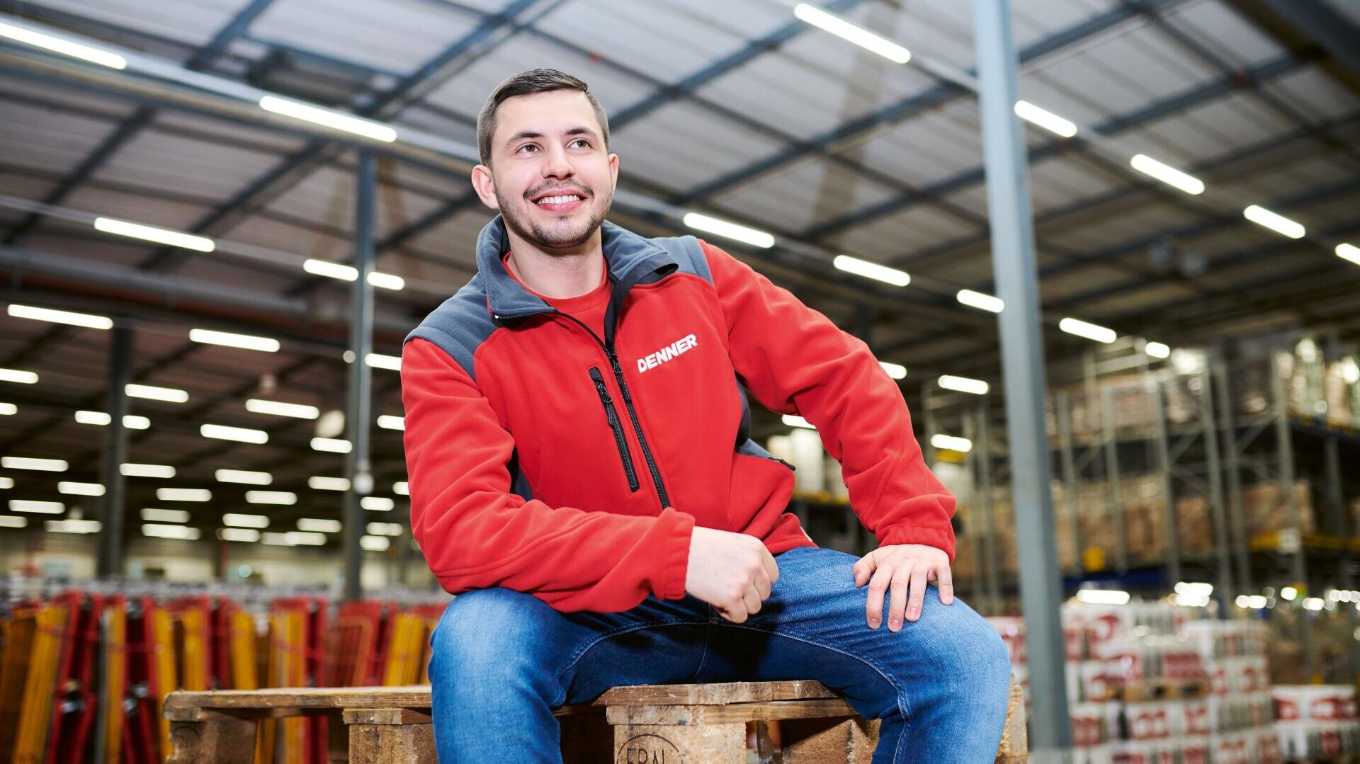 Ein lächelnder Mann in einer roten Jacke sitzt auf einer Holzpalette in einem hell erleuchteten Lagerhaus, in dem im Hintergrund Regale aufgebaut sind.
