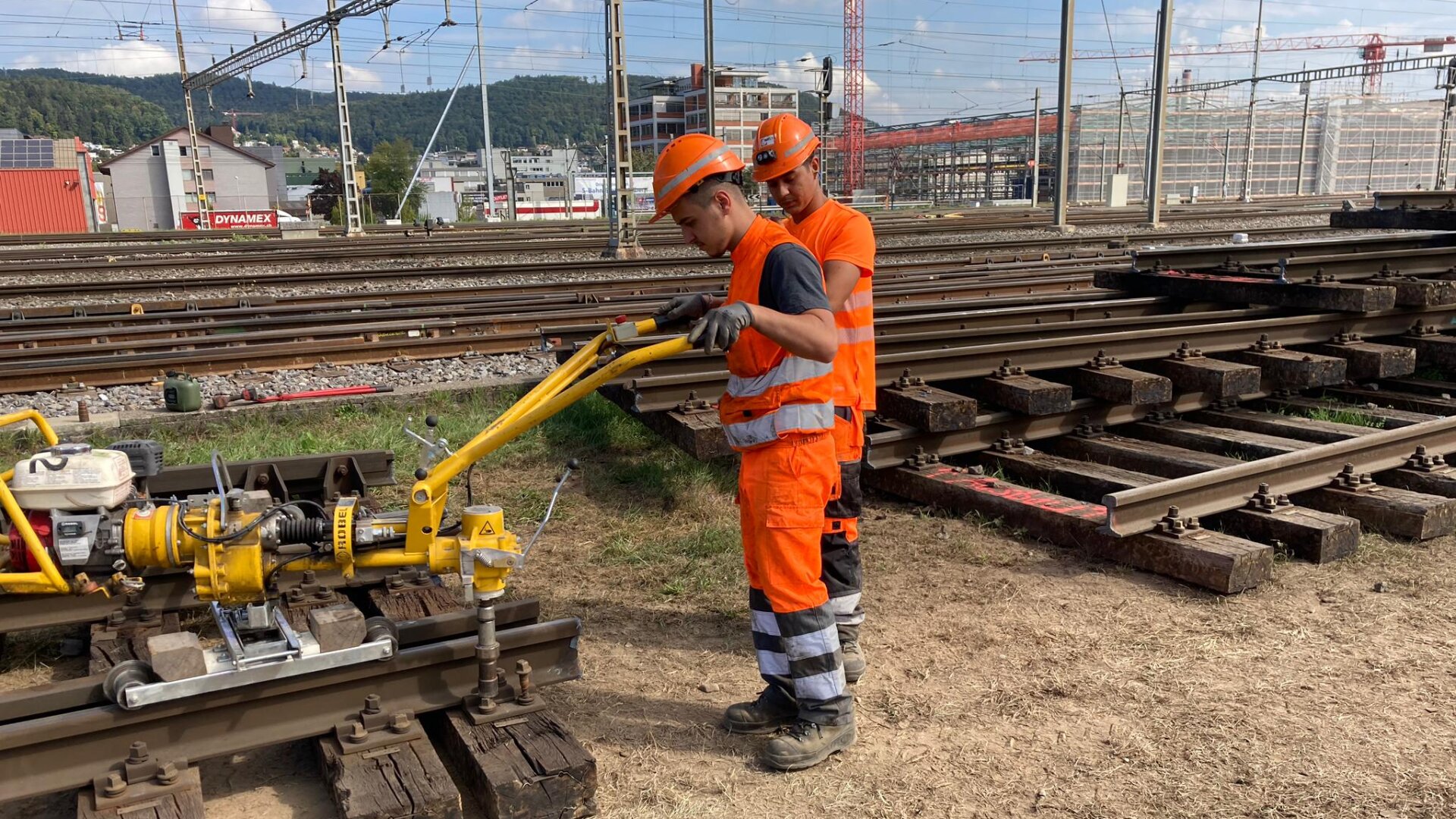 Zwei Eisenbahner in orangefarbener Schutzkleidung bedienen eine gelbe Gleisinstandhaltungsmaschine neben den Bahngleisen auf einer Baustelle, mit Gebäuden und Infrastruktur im Hintergrund.