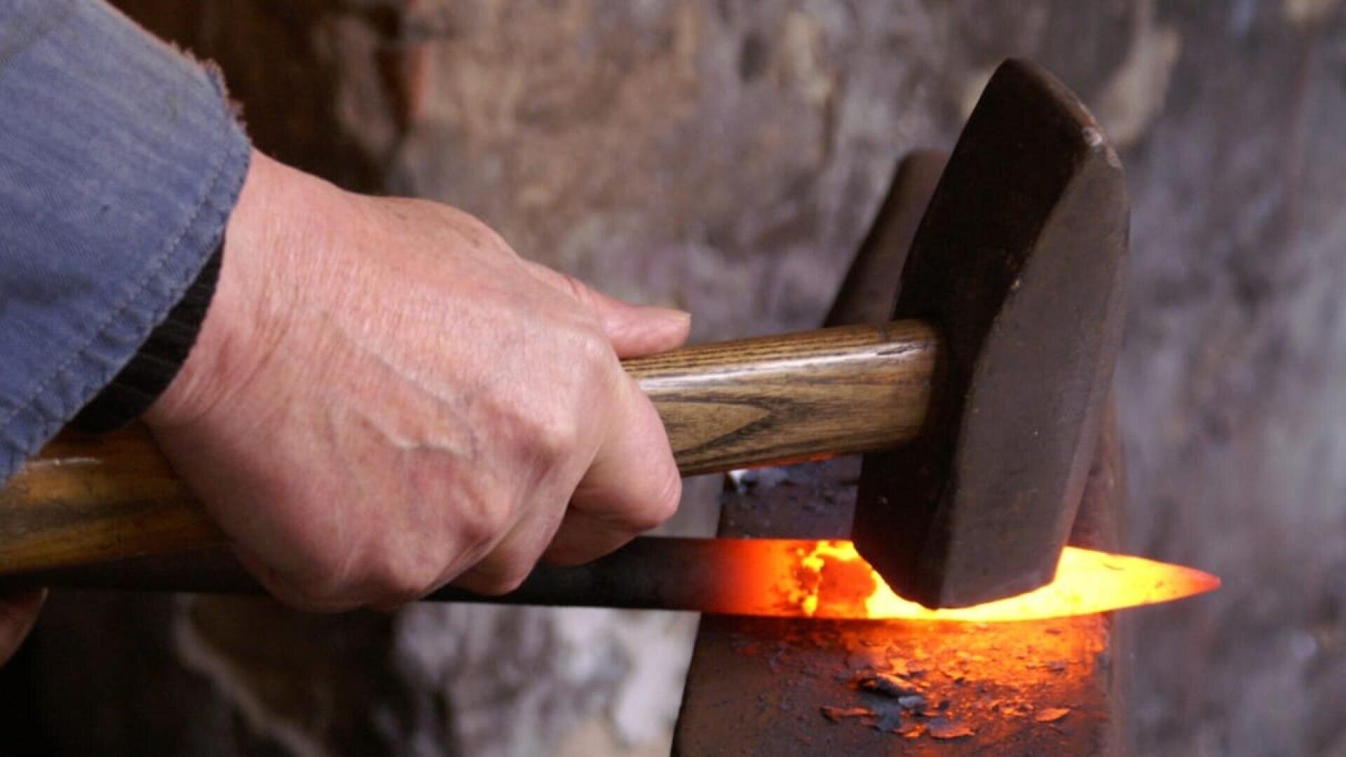 Eine Person schmiedet mit einem Hammer ein glühendes, erhitztes Metallstück auf einem Amboss, wahrscheinlich in einer Schmiedewerkstatt.