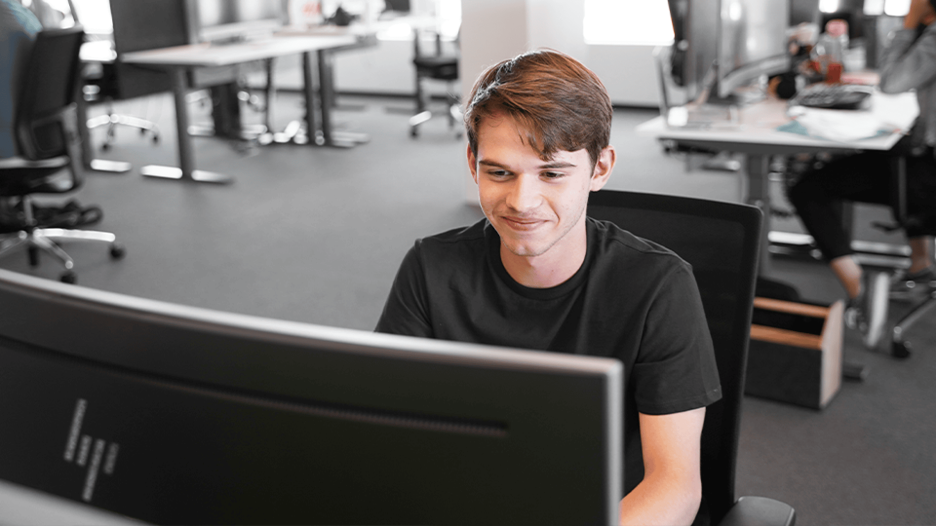 Ein junger Mann mit braunen Haaren, der ein schwarzes T-Shirt trägt, lächelt, während er an einem Computer in einem modernen Großraumbüro arbeitet. Im Hintergrund sind weitere Schreibtische, Stühle und Menschen zu sehen.