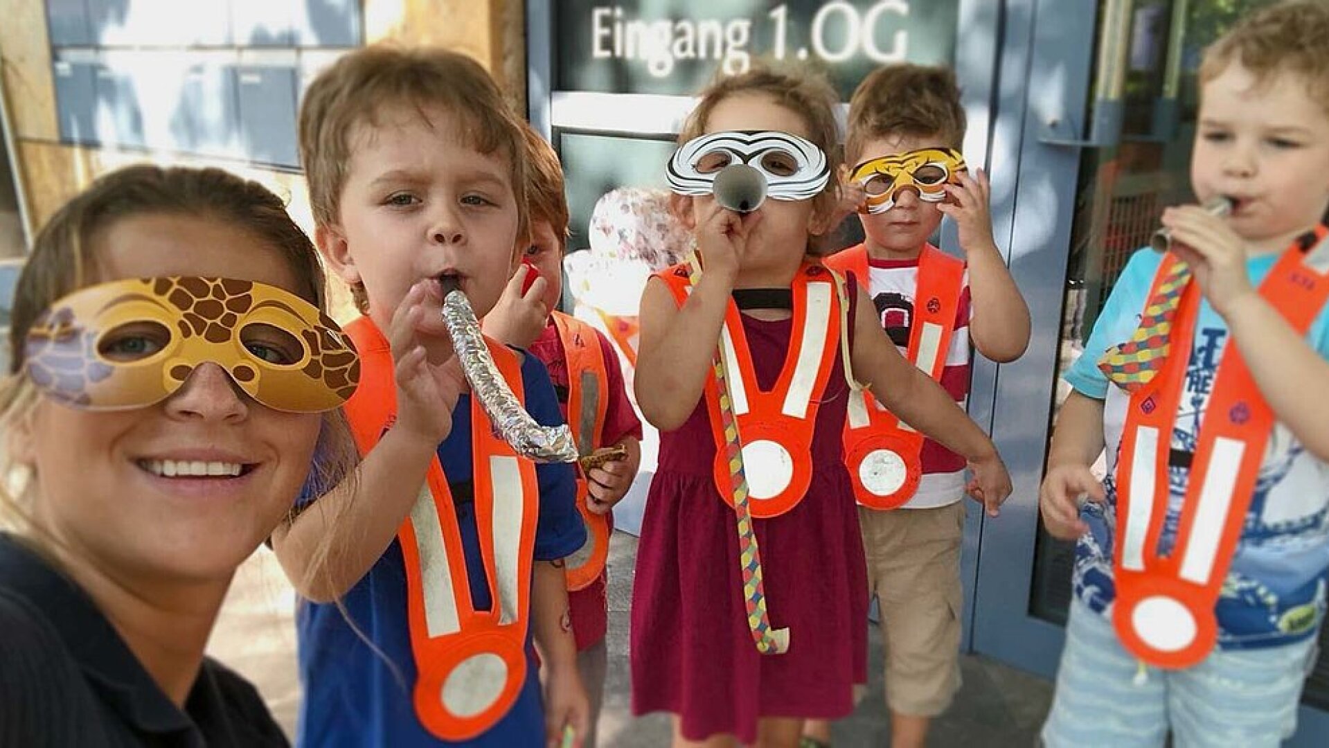 Eine Frau und fünf Kinder mit Tiermasken, orangefarbenen Sicherheitswesten und Partybläsern stehen vor einem Gebäude, lächeln und haben Spaß miteinander.