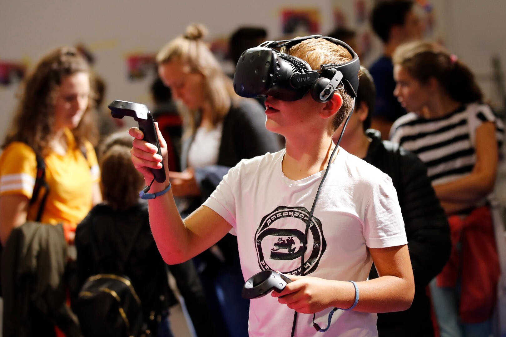 Ein Junge mit einem Virtual-Reality-Headset und Controllern in der Hand taucht in ein VR-Erlebnis ein, umgeben von anderen Menschen in einer belebten Innenumgebung.